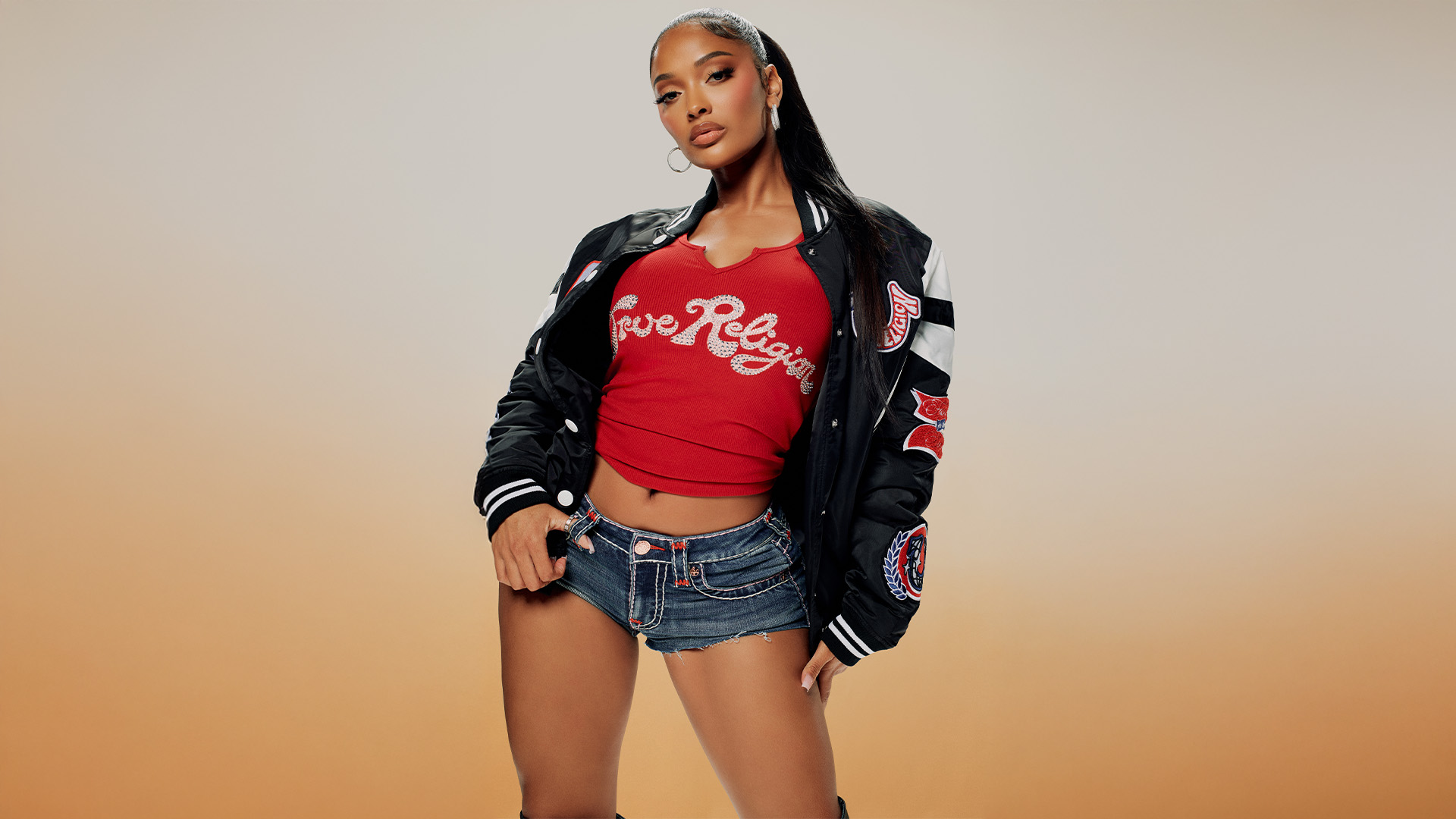 Woman wearing a red true religion tee, true religion jean shorts, and a black and white true religion varsity jacket .