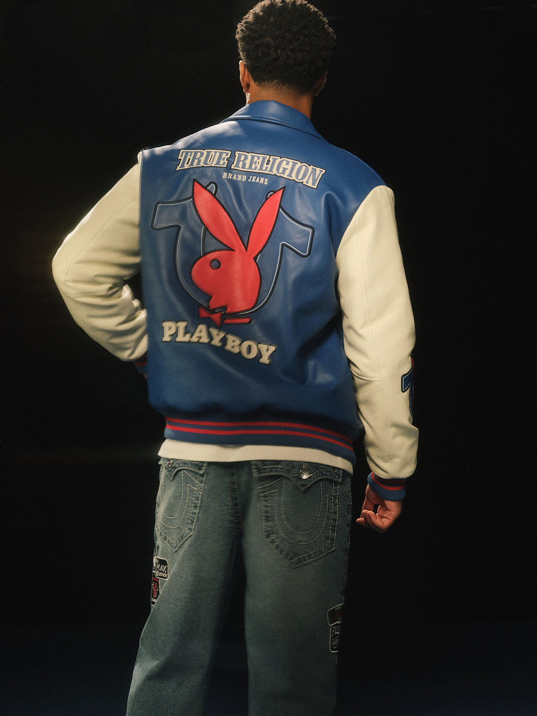 Model wearing a blue and white varsity jacket featuring True Religion and Playboy branding, including a prominent red Playboy bunny logo, paired with relaxed-fit jeans adorned with patch details, set against a dark background.