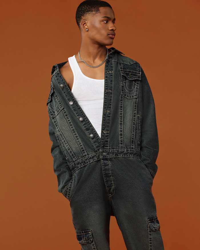 Man wears gray true religion coveralls and a white tank top underneath.