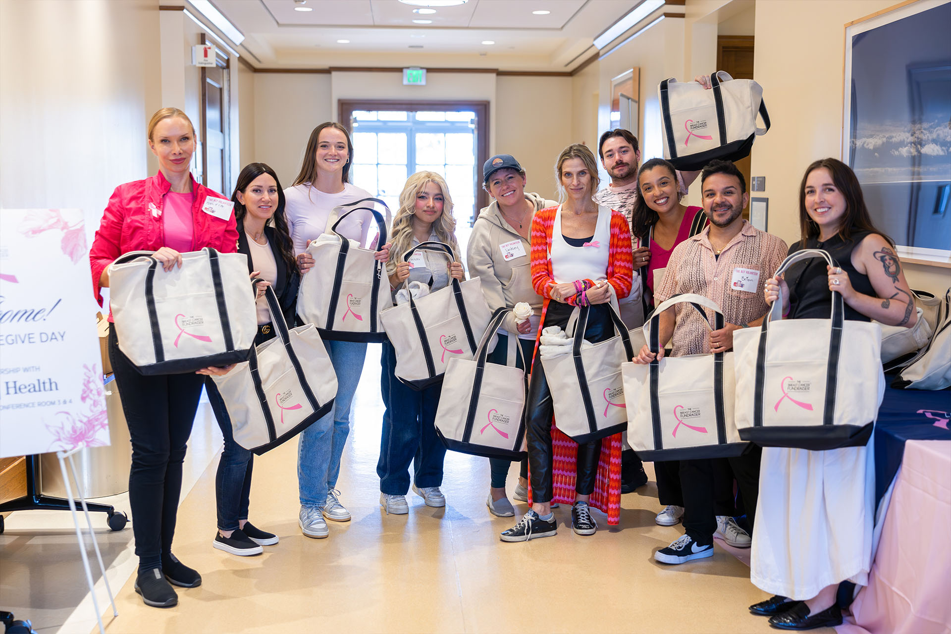 Group of True Religion and Breast Cancer Awareness volunteers indoors holding large tote bags with breast cancer awareness ribbons, smiling for the camera during a community event.