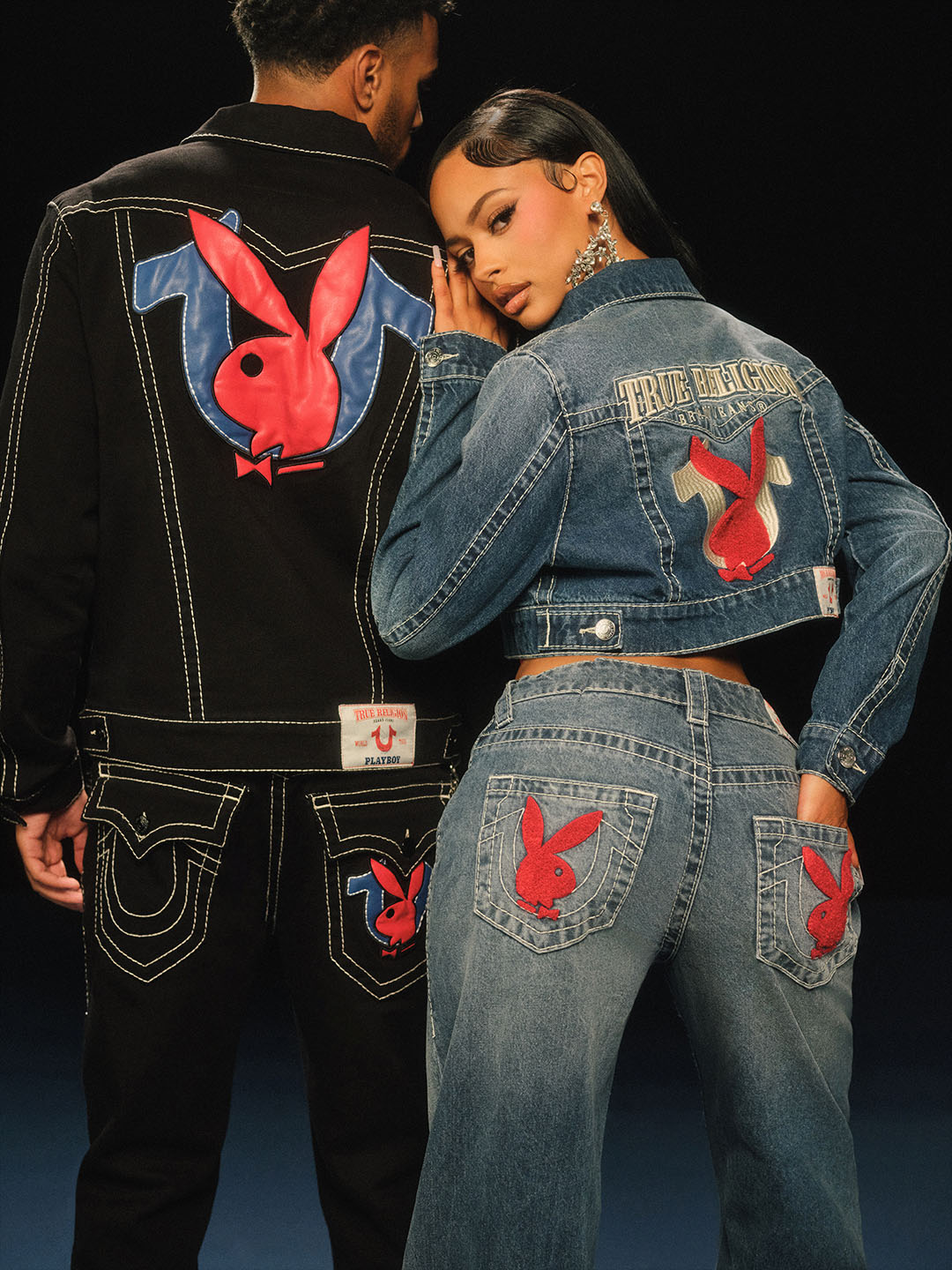 Two models wearing True Religion and Playboy collaboration denim outfits. The woman sports a cropped denim jacket and jeans with red Playboy bunny logos on the pockets, while the man wears a black denim jacket and jeans featuring bold embroidered Playboy and True Religion branding.