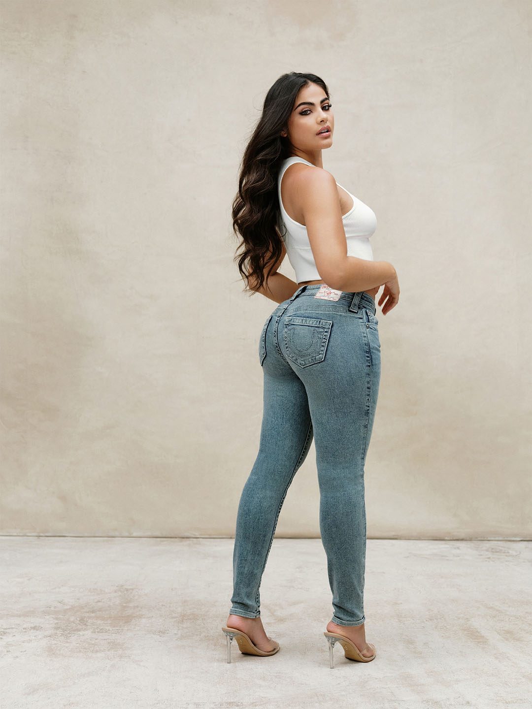 Woman with dark brown hair wearing light wash true religion skinny jeans and a white tank top.