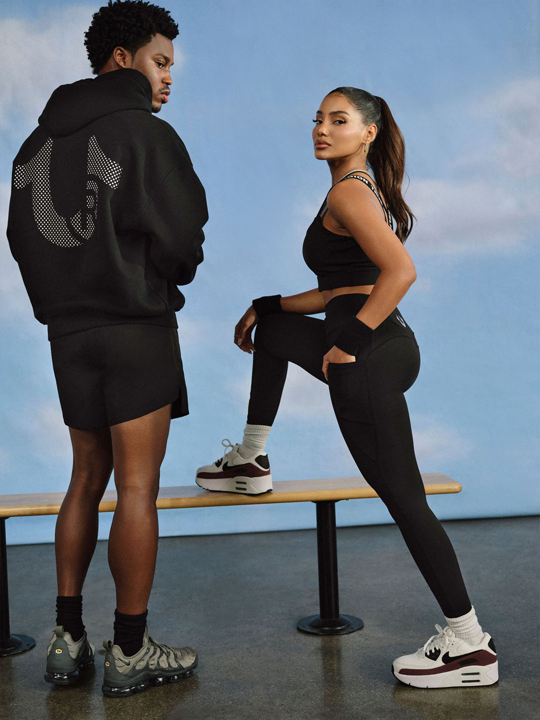 Man and woman wearing black True Religion activewear sets standing with a wooden bench
