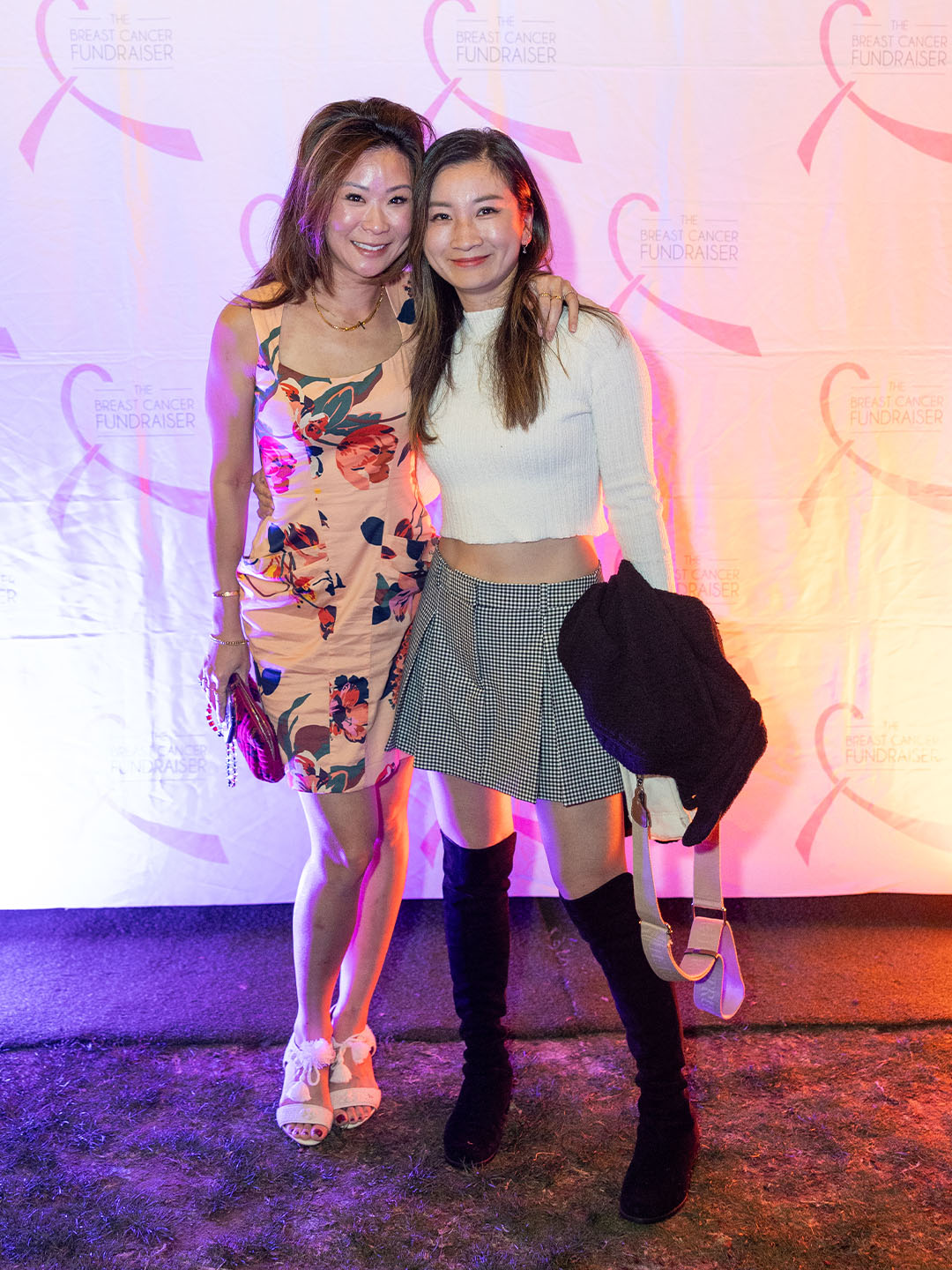 Two True Religion women posing together at a breast cancer fundraiser event, with a pink-themed backdrop featuring the event logo. One is wearing a floral dress with white heels, and the other is dressed in a cropped sweater, plaid skirt, and knee-high black boots.