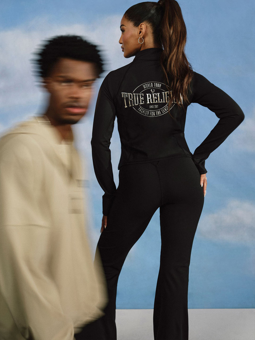 Woman wearing black true religion zip up with white detailing and black yoga pants standing as a man in a beige hoodie walks by.