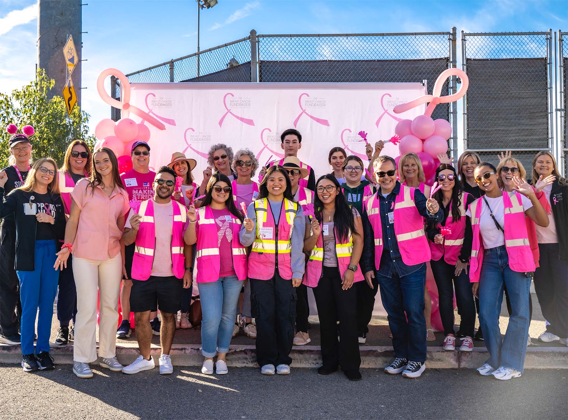 A group of True Religion volunteers and Breast Cancer Awareness volunteers wearing bright pink safety vests and casual clothing pose together in front of a banner featuring pink ribbons at a breast cancer awareness event. The cheerful group stands outdoors on a sunny day, surrounded by balloons and supportive decorations.