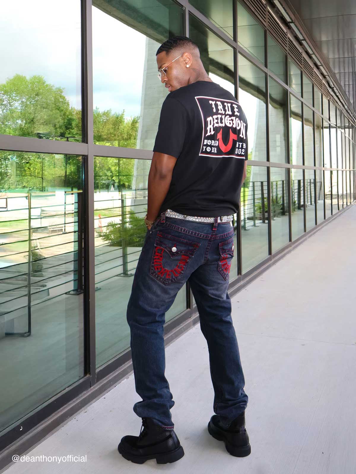Man wears true religion black tee shirt with white lettering and dark wash true religion bootcut jeans with red stitching on pockets