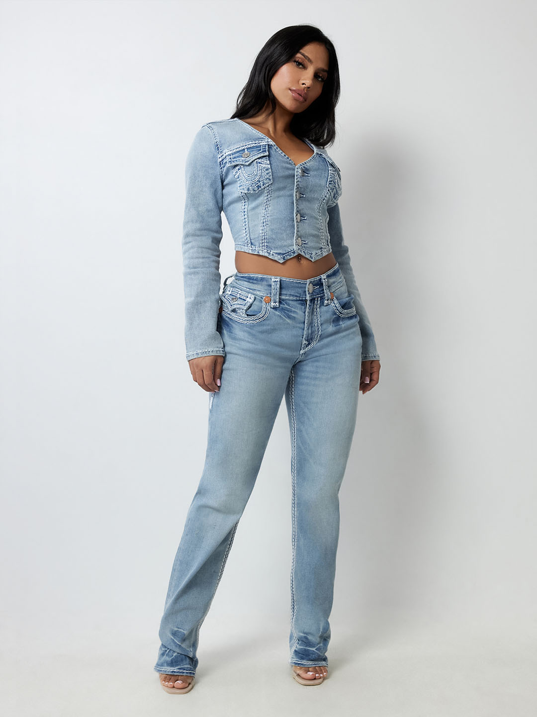 Stylish woman in a light blue denim True Religion set featuring a cropped jacket with button details and matching straight-leg jeans, posing confidently against a plain white background.