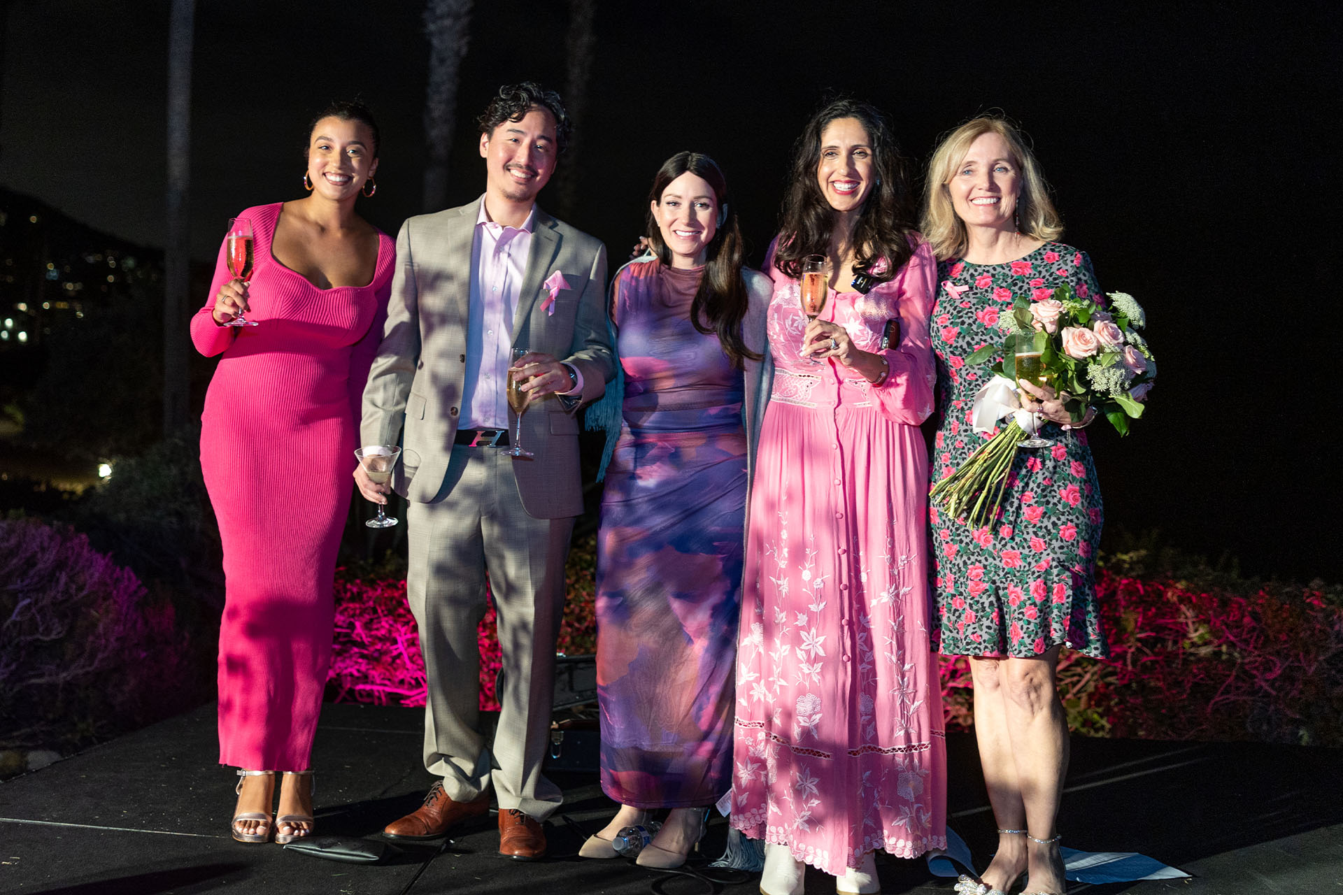 Group of five people posing together at a breast cancer awareness event during the evening. The group dressed in a mix of pink-themed outfits and formalwear. One woman is holding a bouquet of flowers, while others hold champagne or cocktail glasses, with a scenic outdoor nighttime background.