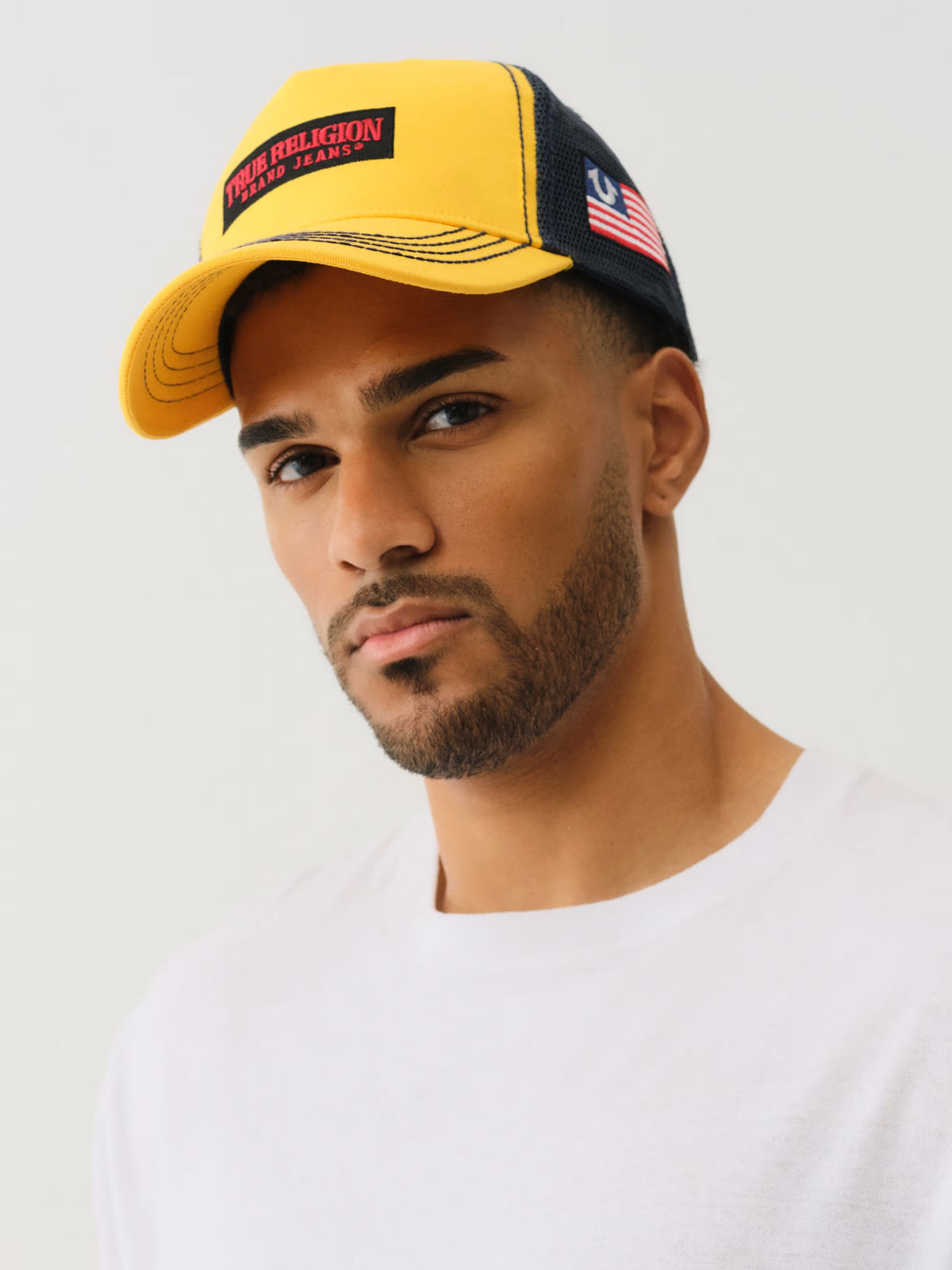 Man wears a yellow true religion trucker hat with red and black detailing, and an american flag stitched on the side