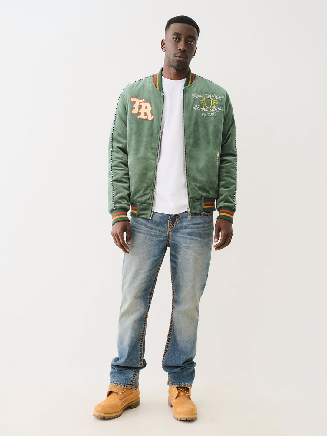 Model wearing a green True Religion varsity-style jacket with embroidered details, paired with a white t-shirt, light wash jeans, and tan work boots, showcasing a casual yet polished streetwear look.