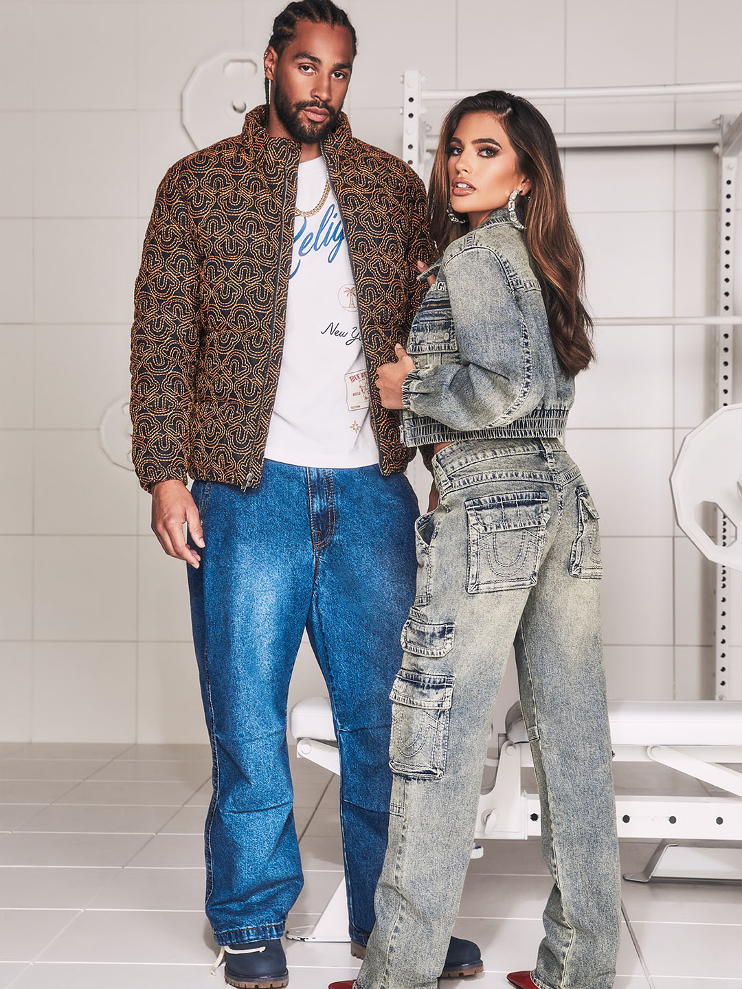 A man wearing true religion blue jeans and a bomber jacket standing next to a woman wearing gray true religion jeans and a jean jacket.