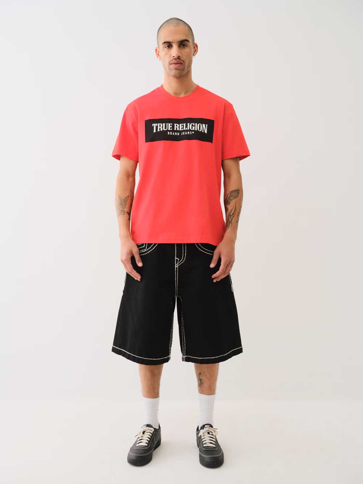Man wears a true religion red tee with black and white logo and true religion black cargo shorts with white stitching