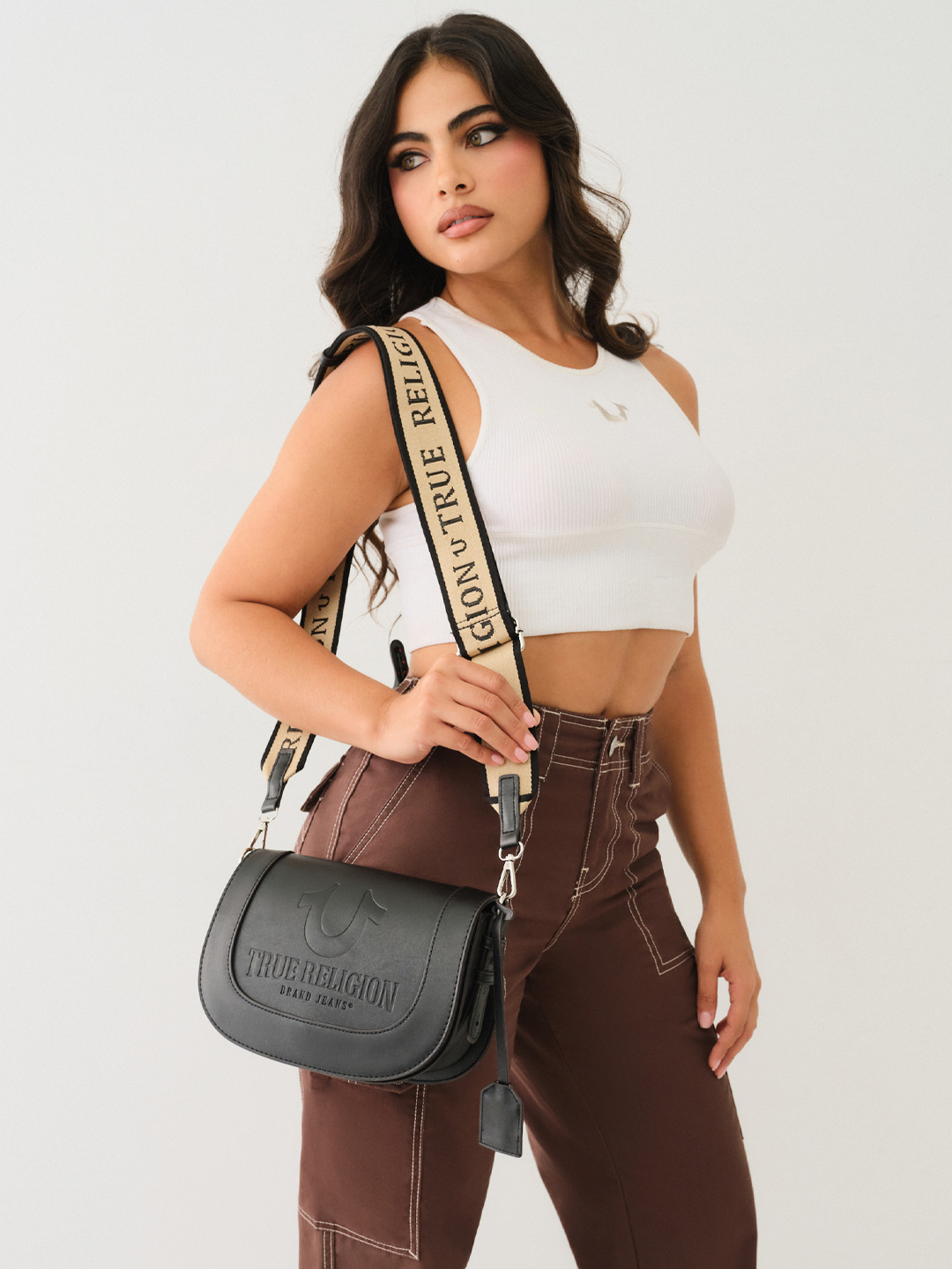 Woman wears a white tank top, brown pants, and the true religion stitched horseshoe flap crossbody bag