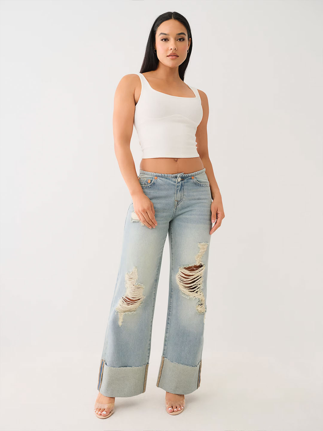 Woman wearing a white tank top, light wash ripped true religion jeans with a frayed hem, and nude heels.
