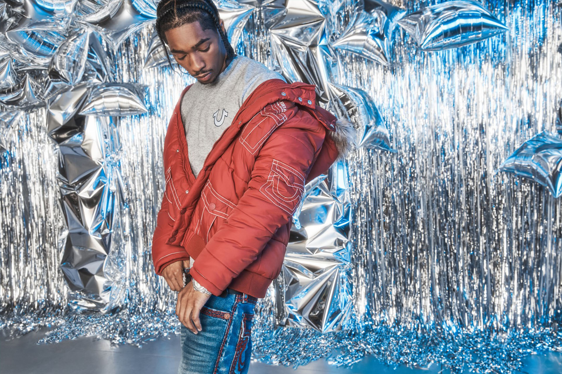 Man wearing a true religion red puffer jacket, gray t-shirt, and true religion blue jeans with red stitching standing in front of a background of silver balloons and streamers.