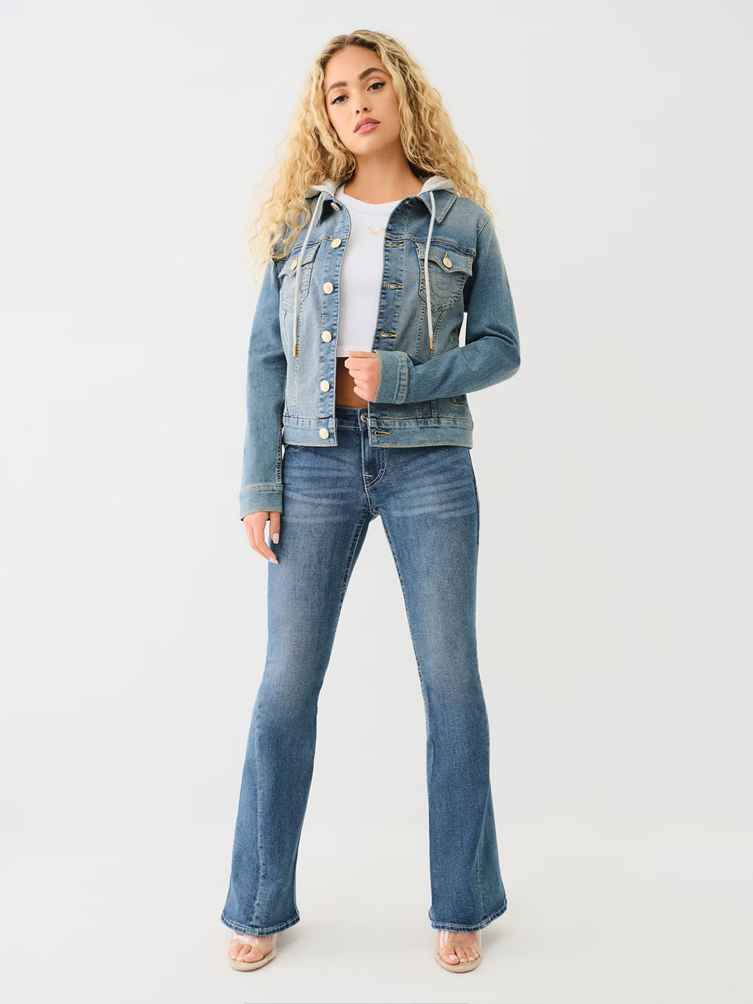 Woman wearing a light wash jean jacket with a hood, a white t shirt, and blue true religion jeans.