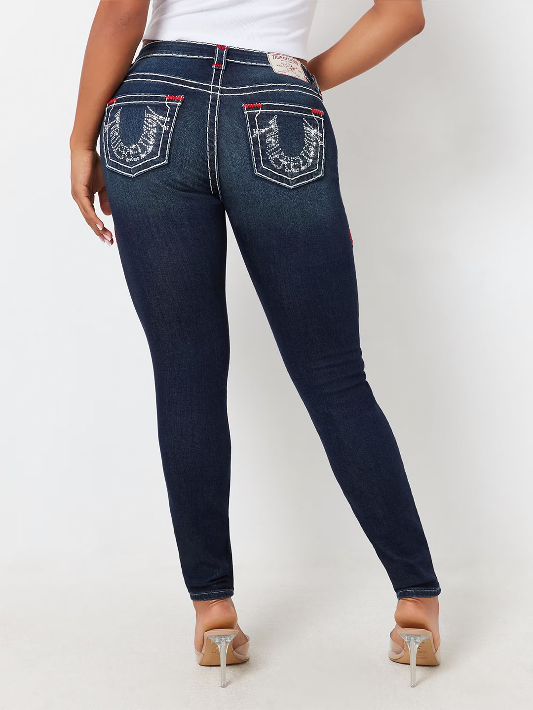 Back view of a model wearing dark wash True Religion skinny jeans featuring signature horseshoe embroidery on the pockets with white stitching and red accents, paired with a white top and clear high heels.