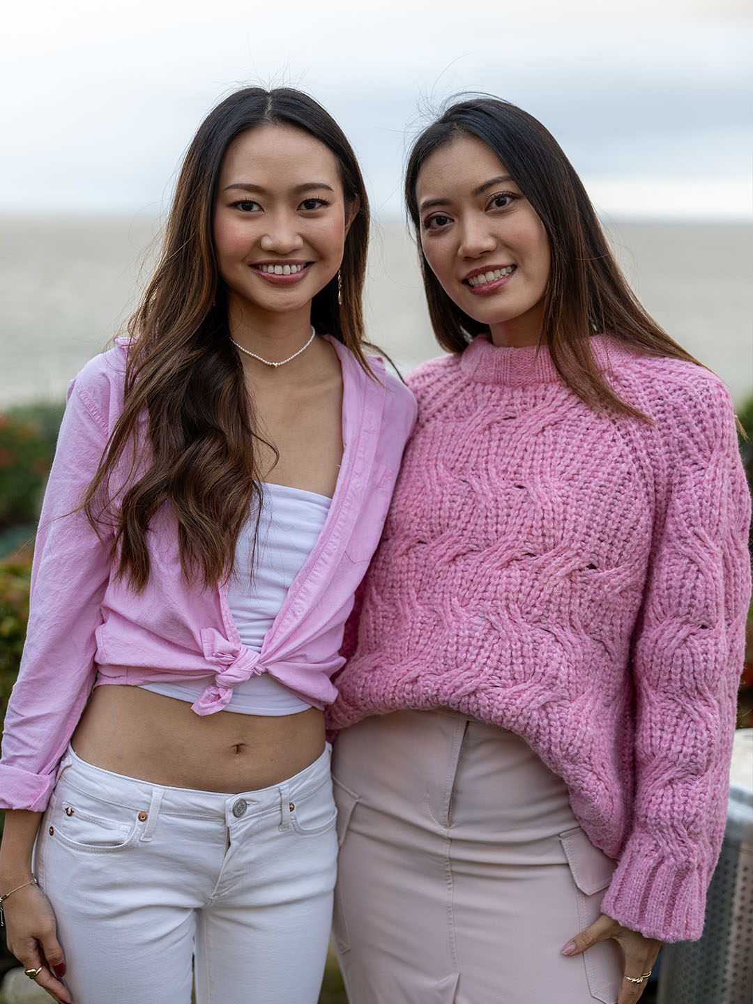Two True Religion employee women smiling and posing outdoors with a serene ocean background. One is wearing a light pink cropped blouse paired with white pants, while the other is dressed in a cozy pink cable-knit sweater and light-colored skirt.
