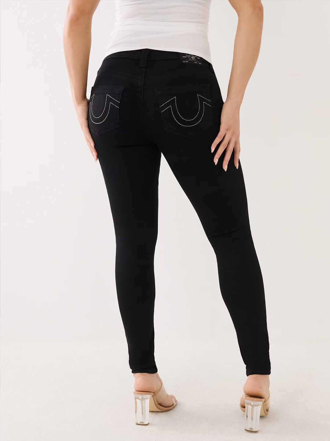 Woman wearing true religion dark wash skinny jeans with white stitching and clear high heels.