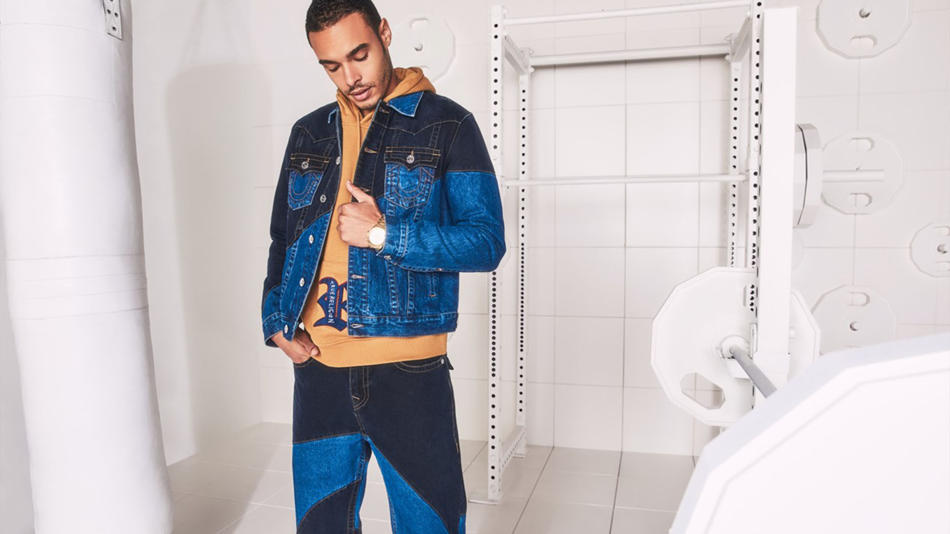 Man poses against a white industrial background wearing a denim jacket, brown sweatshirt, and light and dark wash pattern jeans.
