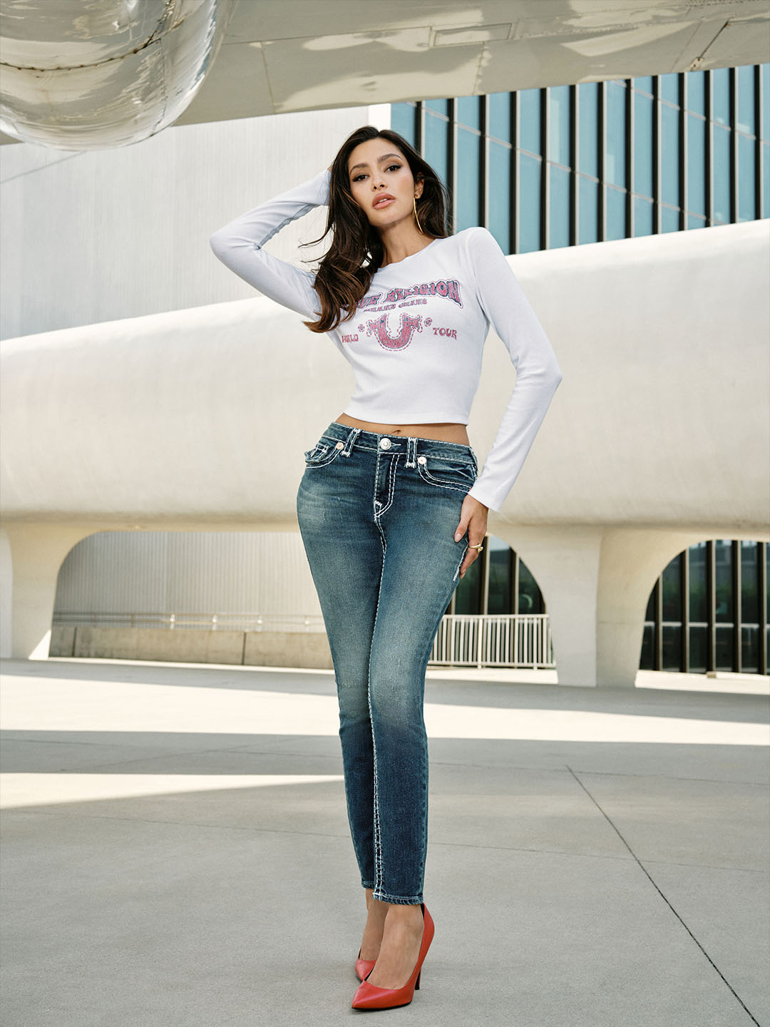 Woman wearing a white long sleeved cropped shirt with gray true religion skinny jeans with white stitching, standing on concrete.
