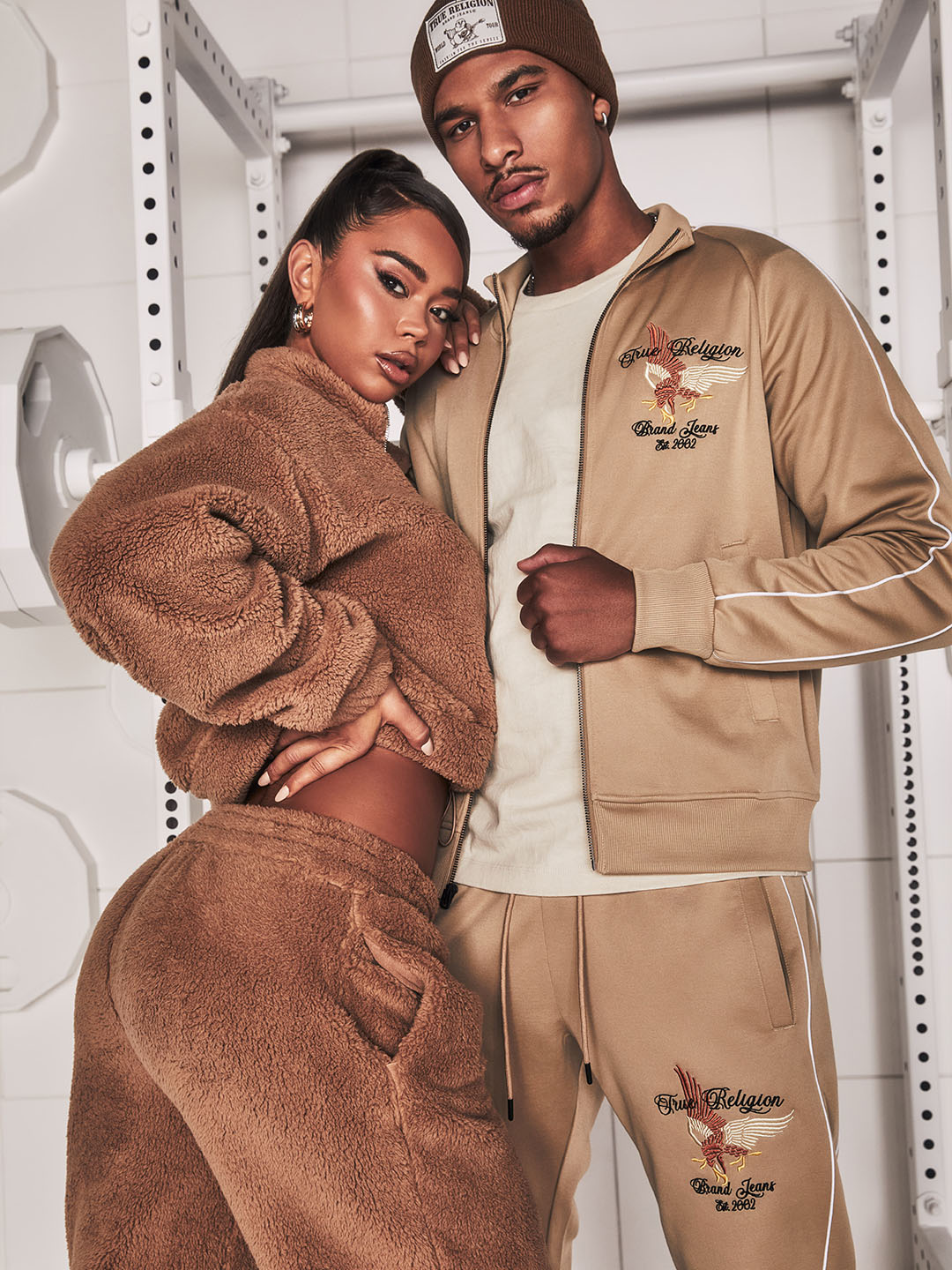 Stylish duo wearing coordinated beige outfits; the woman in a cozy fleece set and the man in a tracksuit with embroidered details, posing confidently in a modern gym-inspired setting.