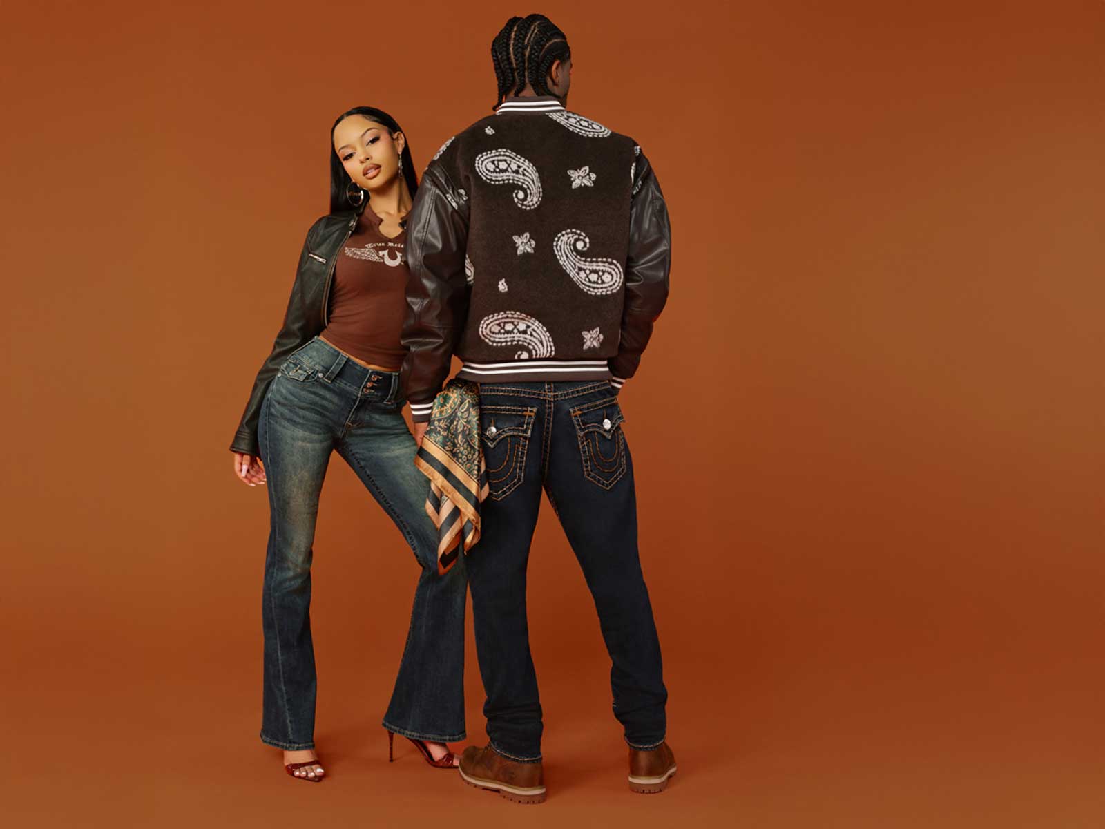 Woman wears true religion brown baby tee with true religion dark wash flared jeans, and man wears true religion brown paisley varsity jacket and true religion dark wash baggy jeans with stitching
