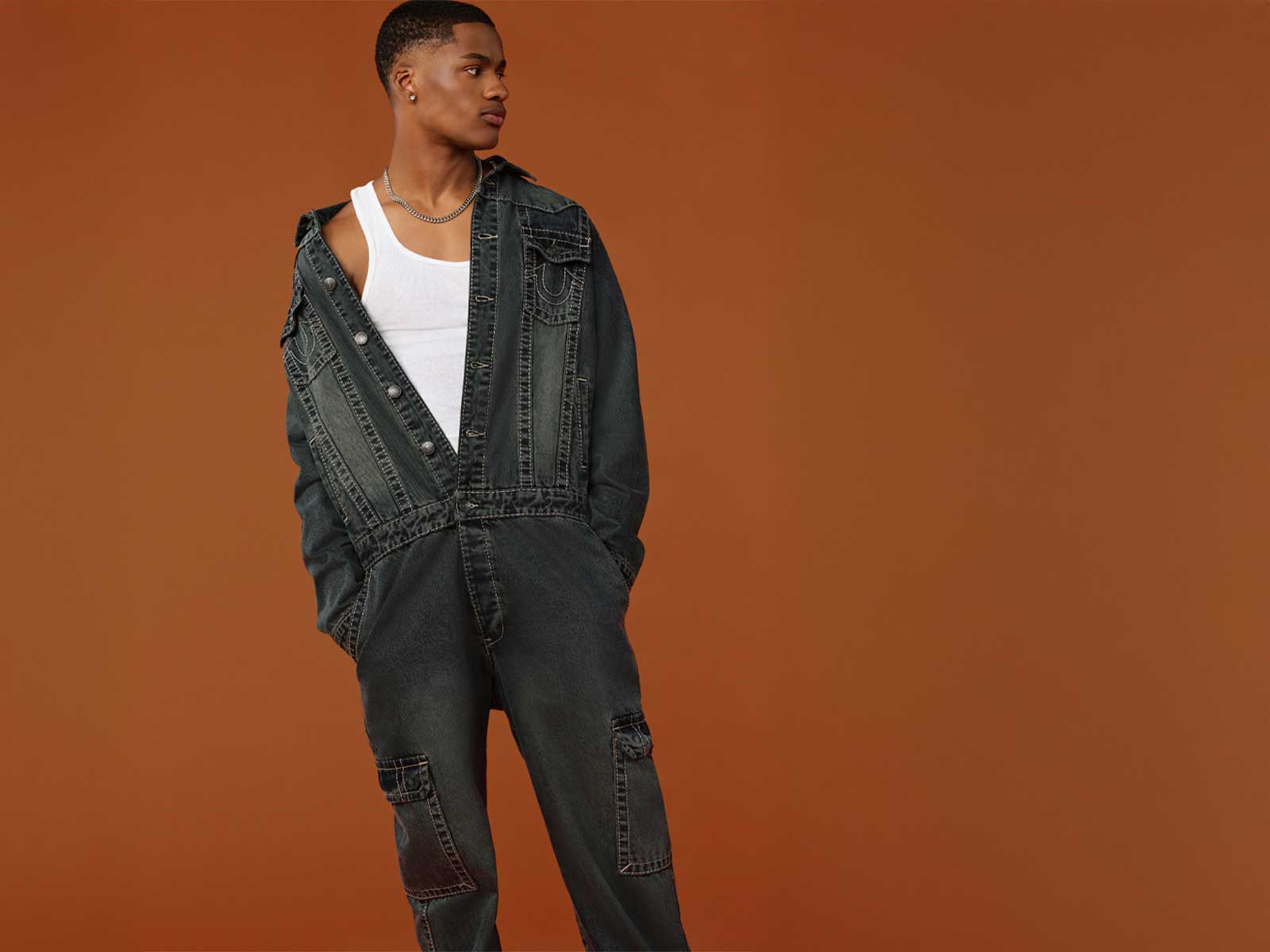 Man wears gray true religion coveralls and a white tank top underneath.