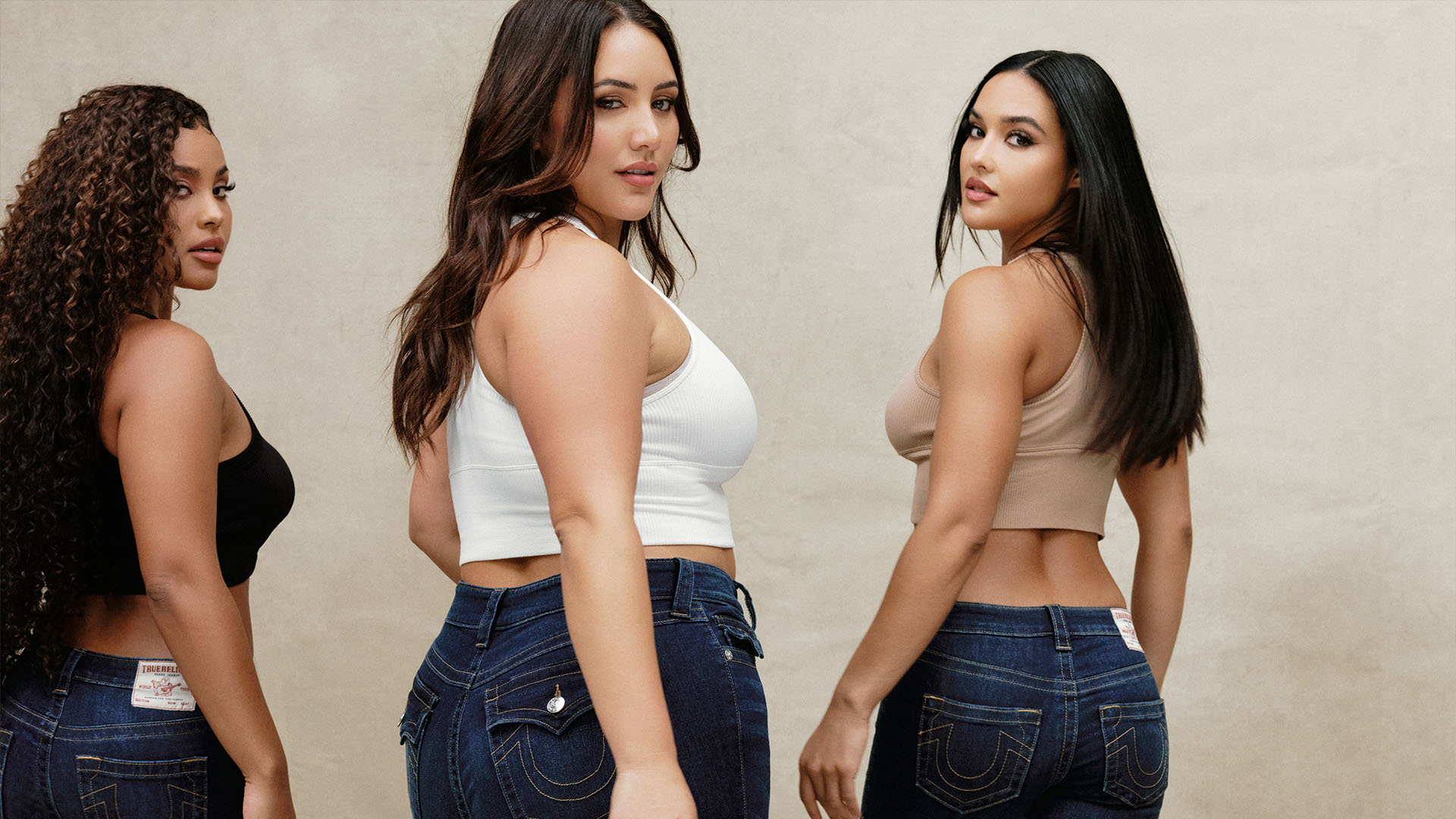 Three women in tank tops wearing different sizes of dark wash true religion jeans.