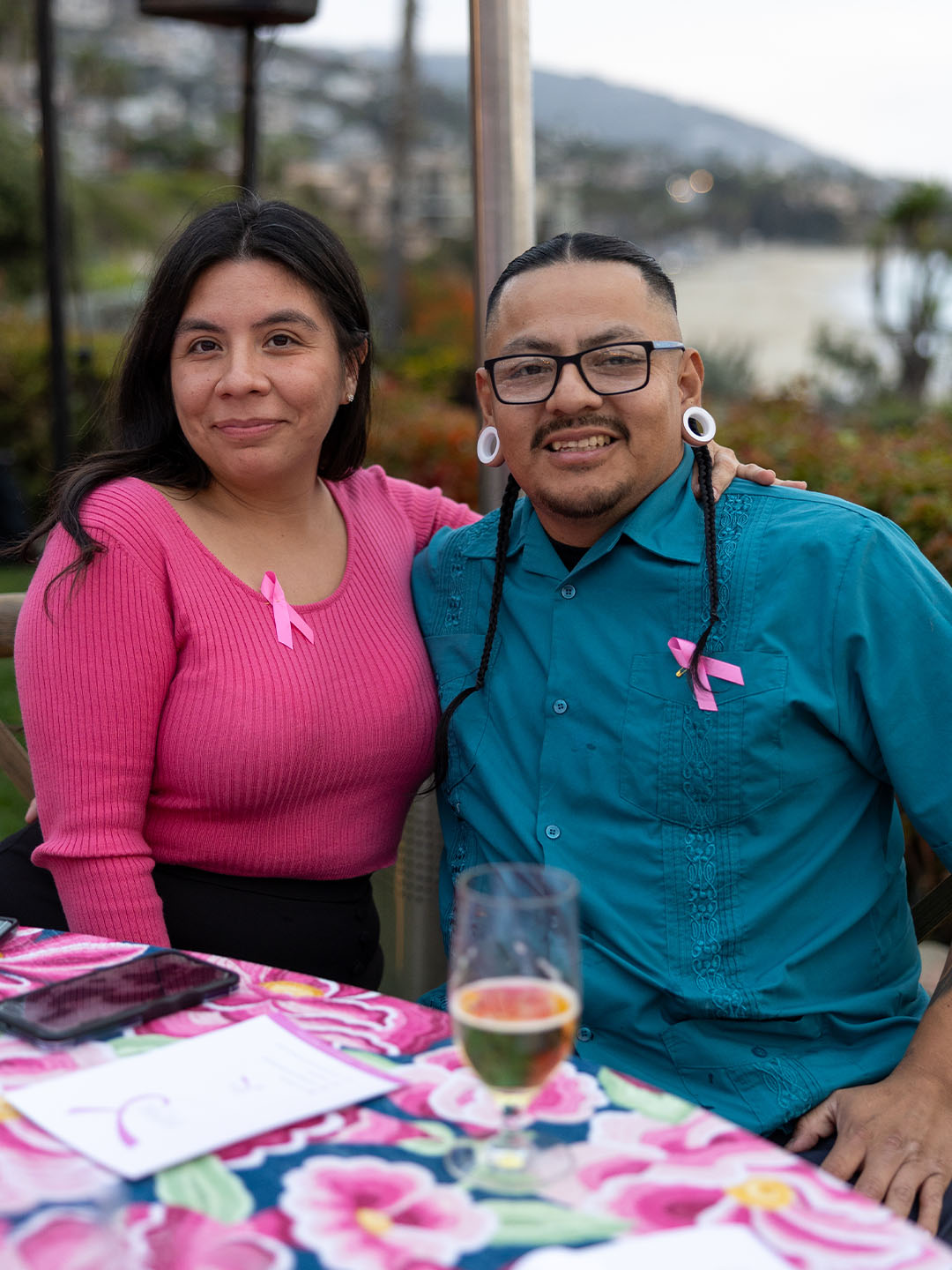 A smiling True Religion couple seated at a floral-patterned table outdoors with a scenic coastal background. The woman is wearing a bright pink top with a breast cancer awareness ribbon, and the man is dressed in a teal shirt with matching ribbon.