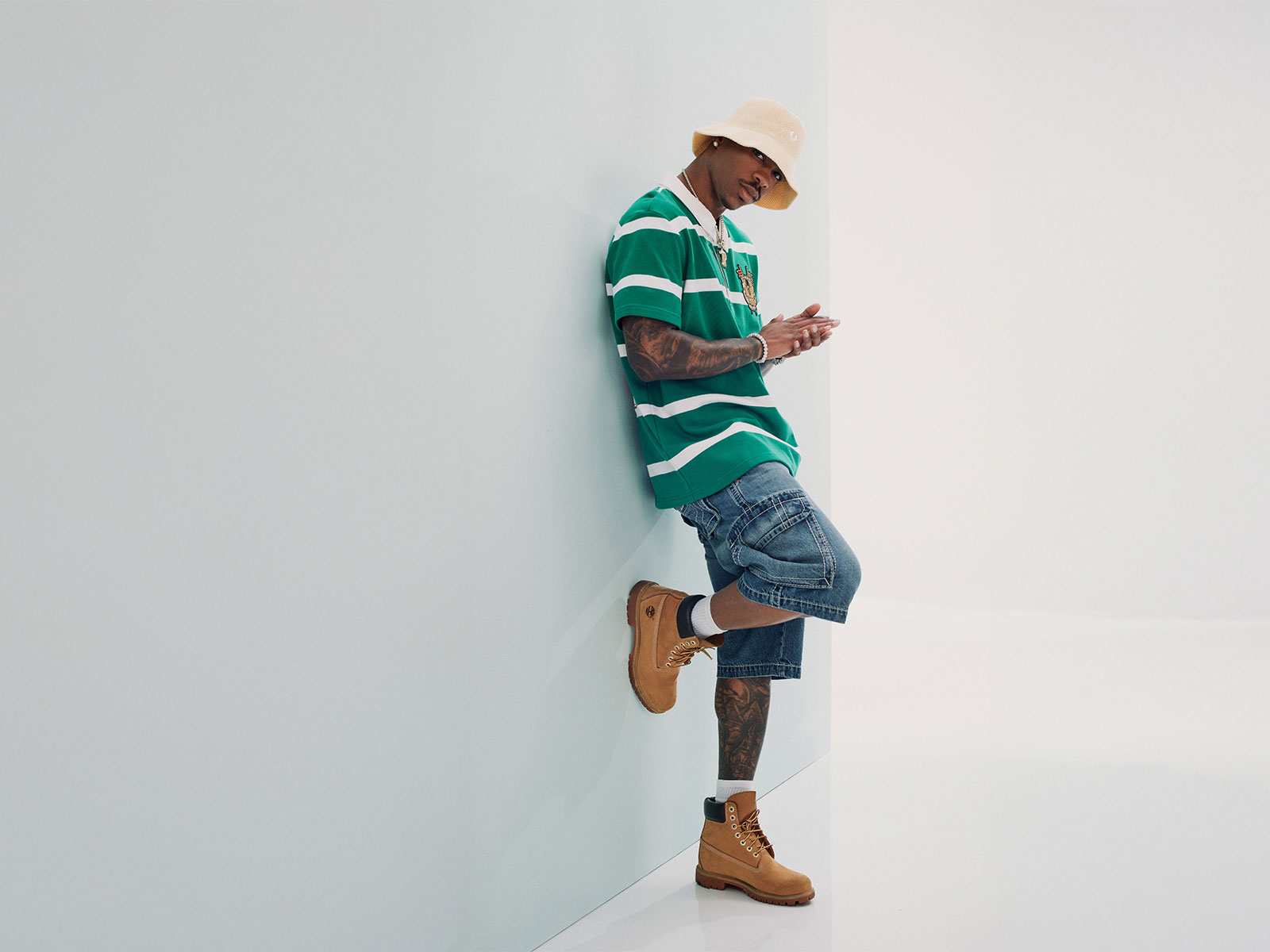 Man leans against a white wall wearing true religion denim shorts, a true religion green and white striped polo tee, and brown boots