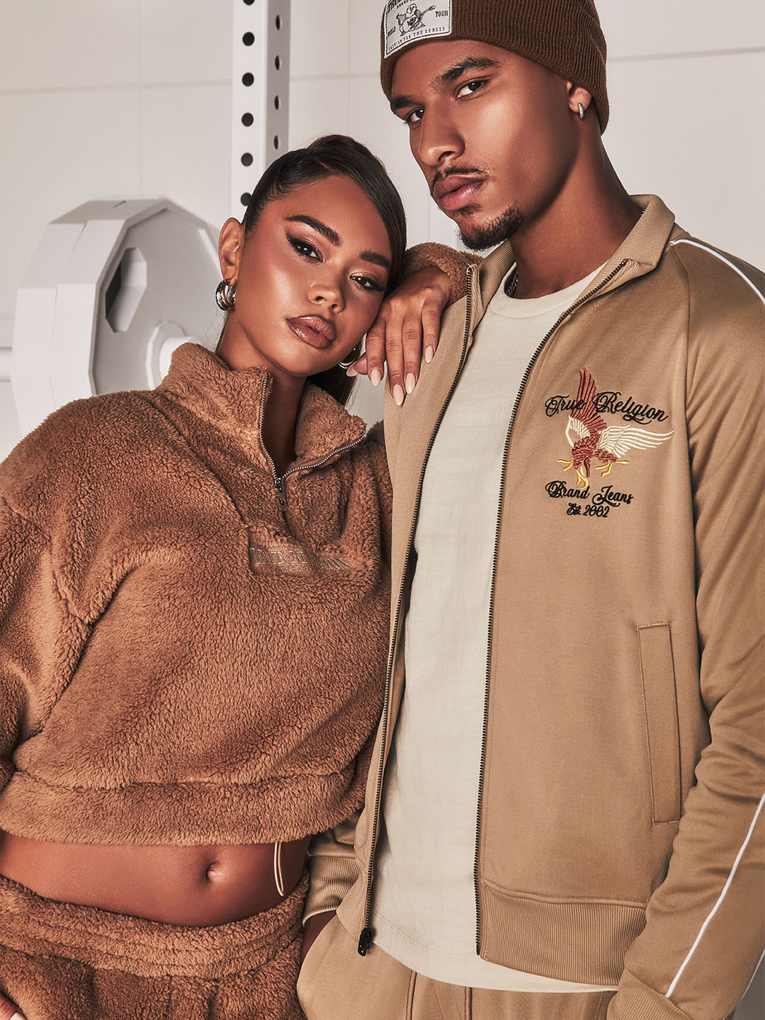 Close-up of a fashionable pair in coordinated beige loungewear; the woman in a fleece crop top and the man in a tracksuit with embroidered accents, exuding confidence in a sleek gym-inspired setting.