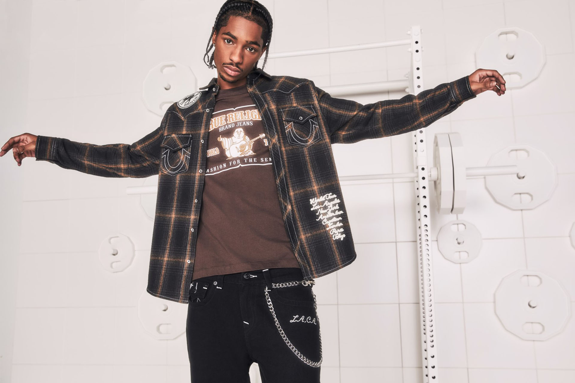 Man posing with arms outstretched against a white background, wearing a brown true religion shirt, a plaid jacket with white horseshow stitching, and black true religion jeans.