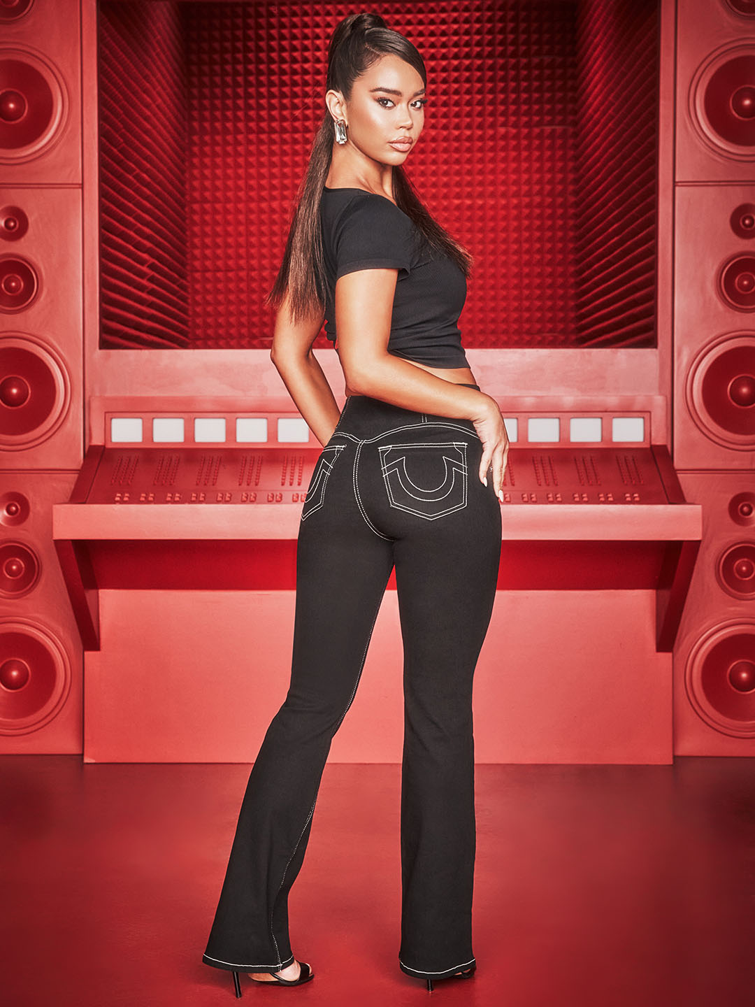 Woman turning towards the camera wearing a balck baby tee and black true religion bootcut jeans with white stitching.