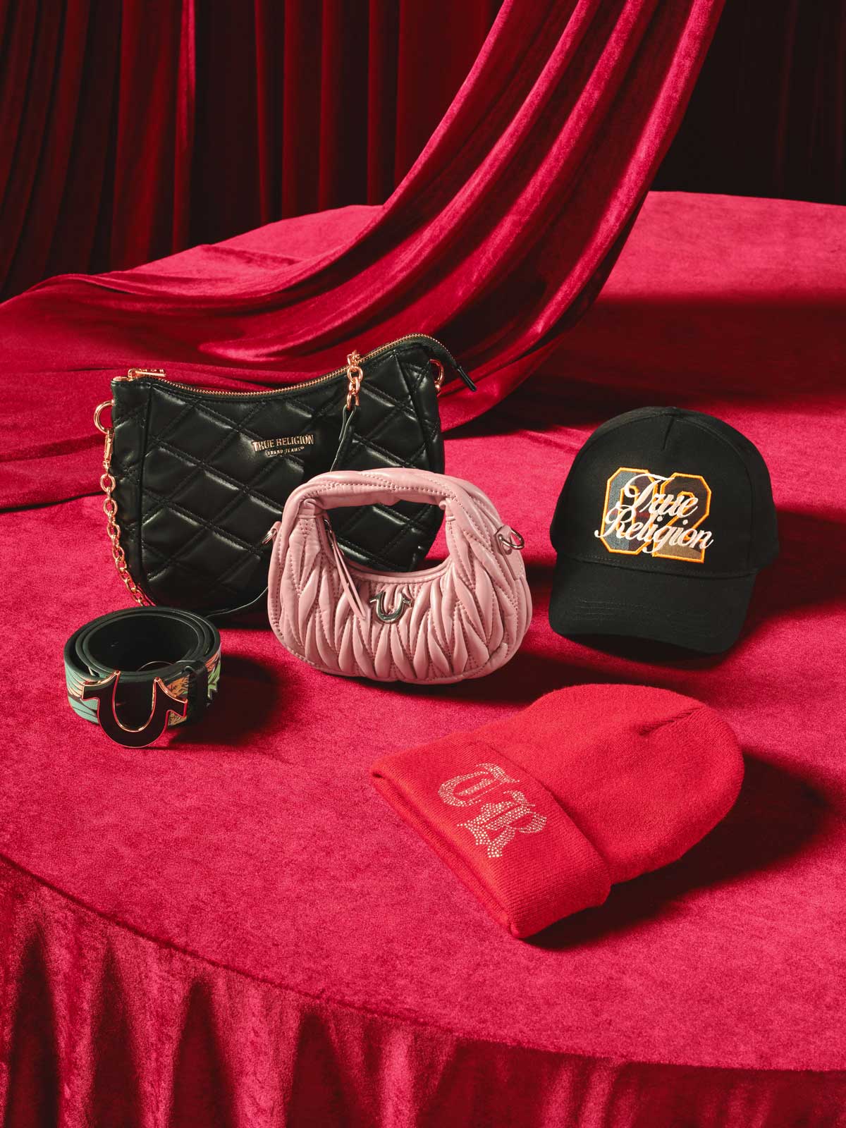 true religion accessories sitting on a table: a black purse, a pink purse, a black belt with horseshoe buckle, a black beanie, and a red trucker hat