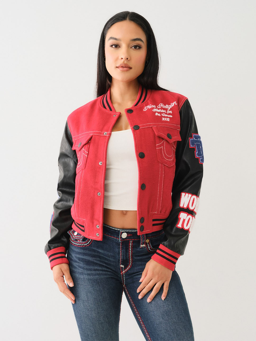 Woman wearing a white tank top, true religion blue jeans with red and white stitching, and a true religion red and black bomber jacket.