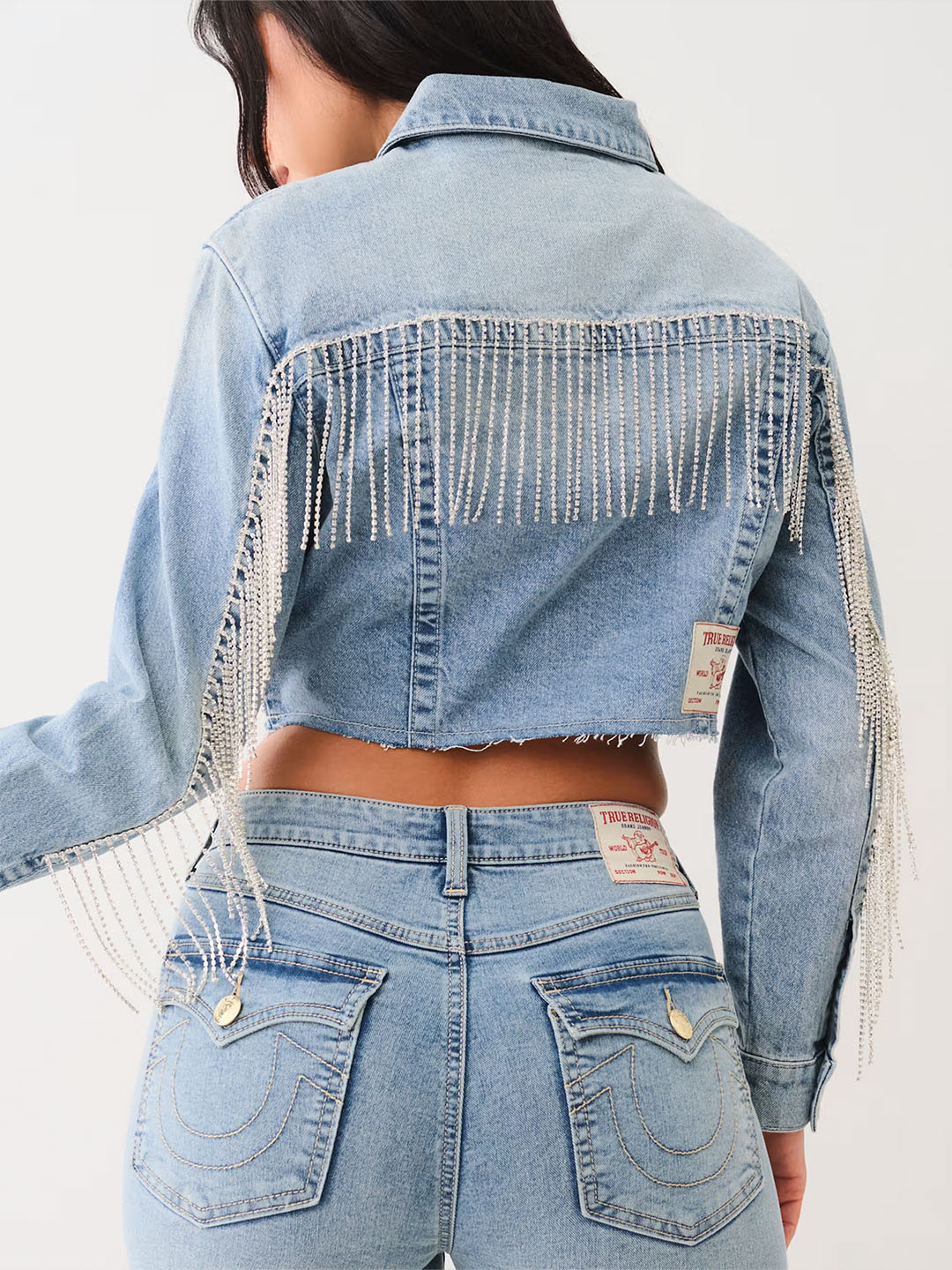 Woman wearing a true religion denim jacket with rhinestone fringe and true religion light wash jeans.