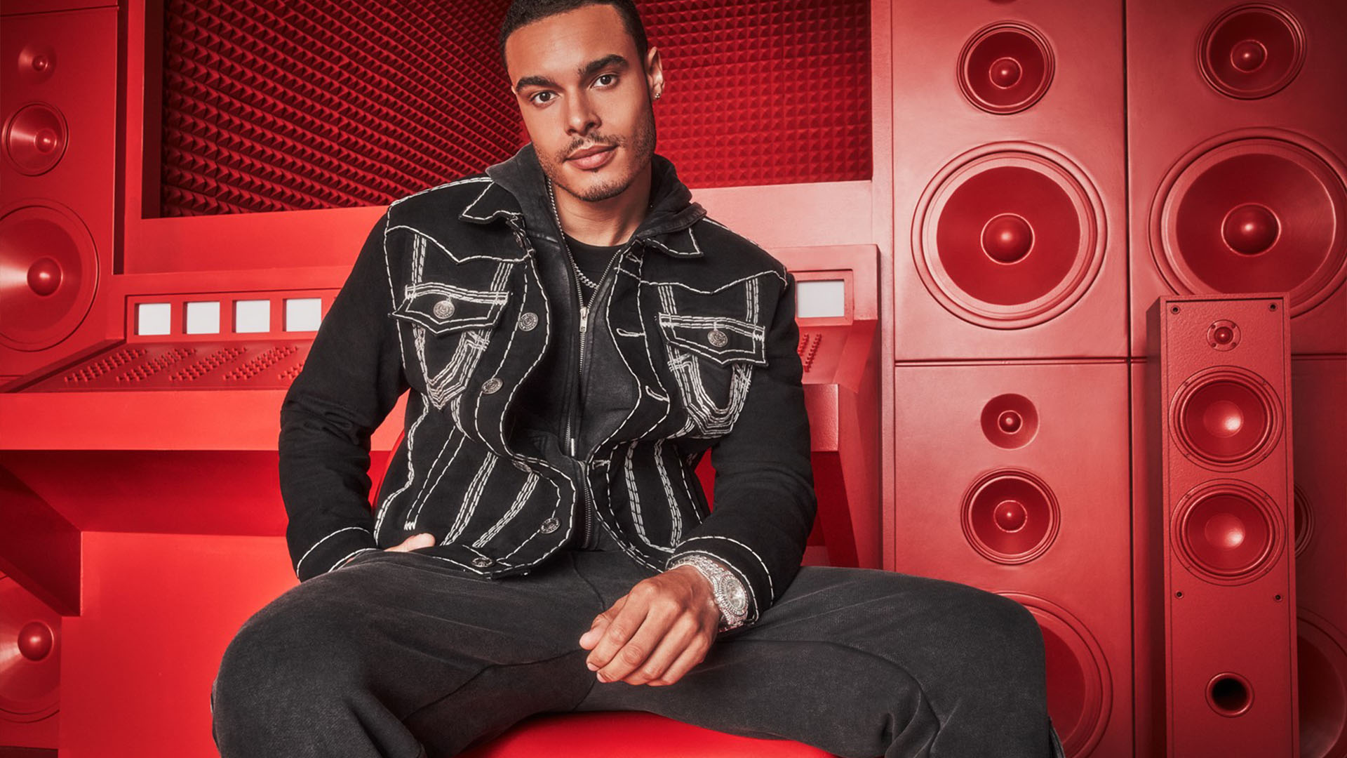Man dressed in a black true religion jean jacket with white stitching and dark wash true religion jeans sits in front of a red background