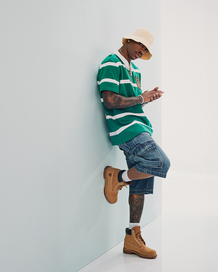 Man leans against a white wall wearing true religion denim shorts, a true religion green and white striped polo tee, and brown boots