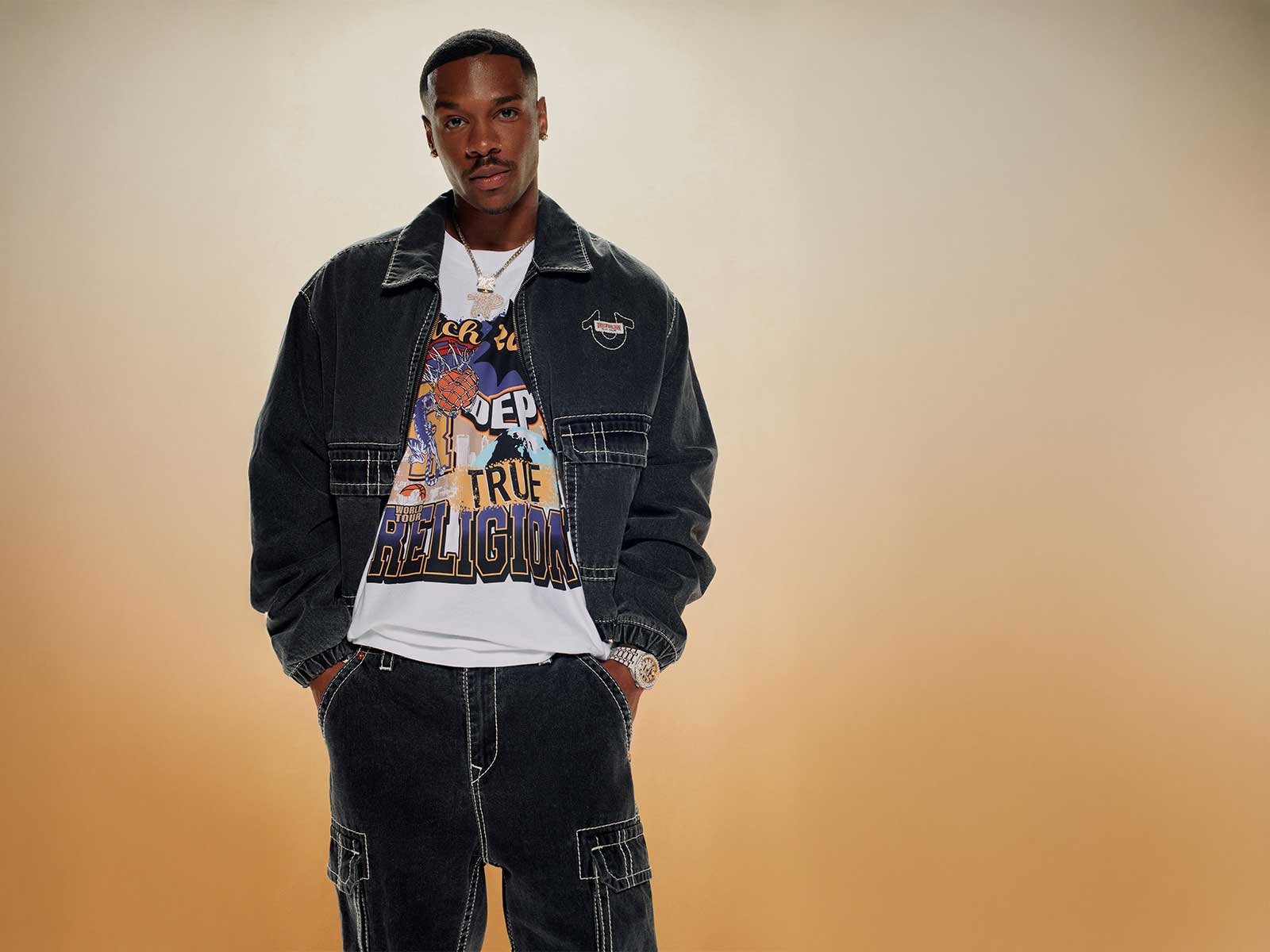 Man wears true religion black cargo pants with white stitching, a black true religion jacket with white stitching, and a white graphic tee