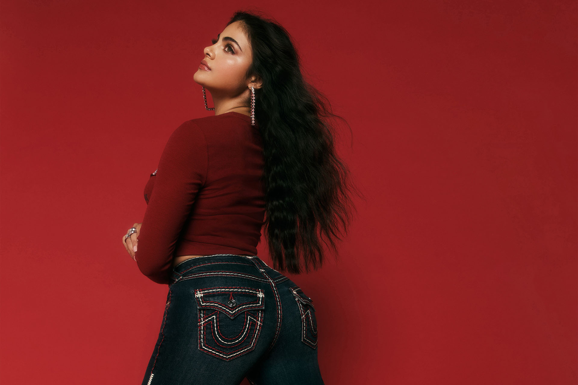Woman that is kneeling, wearing dark wash true religion jeans with white and black stitching against a red background.