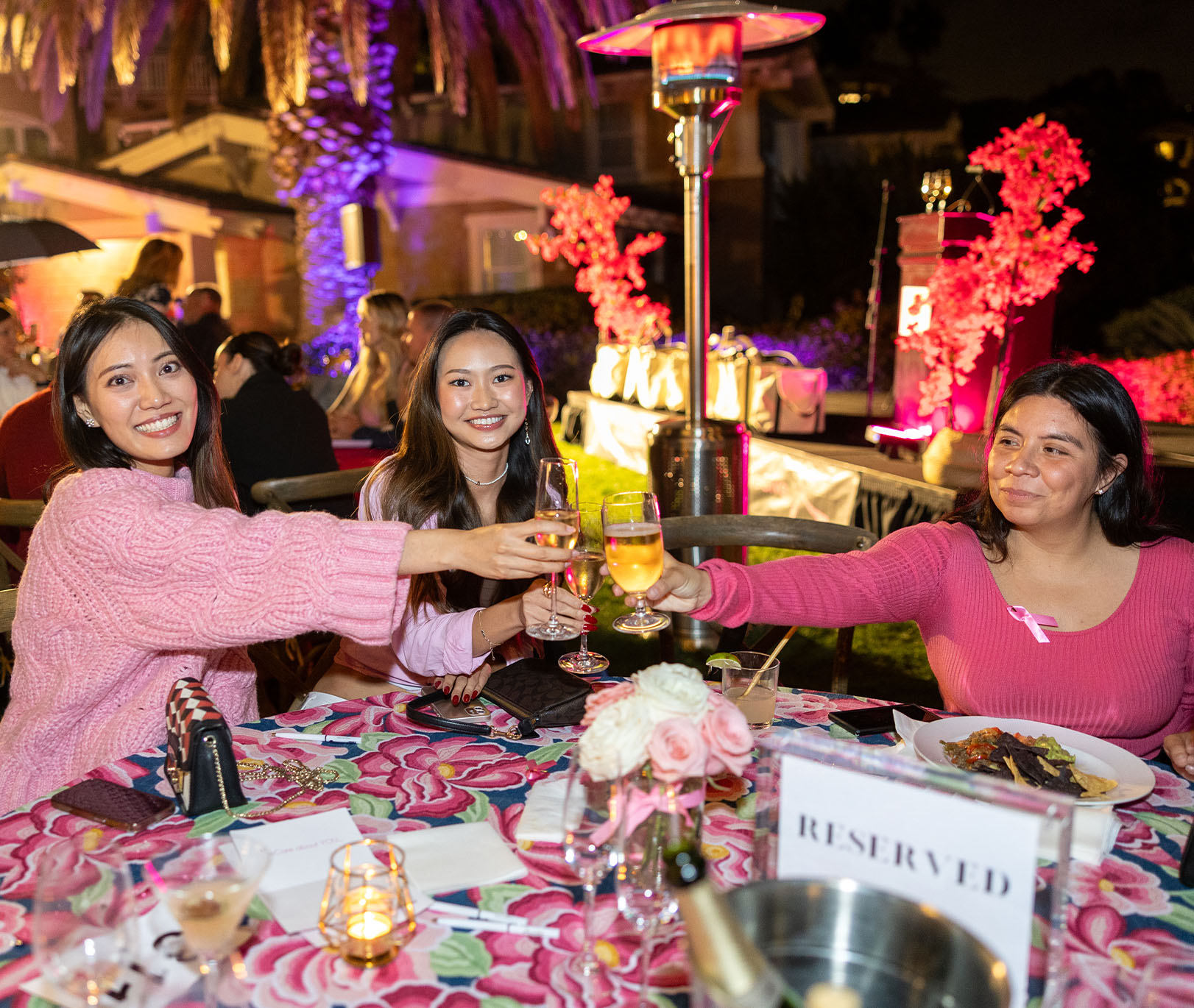 Three True Religion women employees seated at a colorful floral-patterned table outdoors, raising their glasses in a toast during an evening event. The scene is illuminated by festive lights, with a vibrant background featuring decorative trees, heat lamps, and a podium.