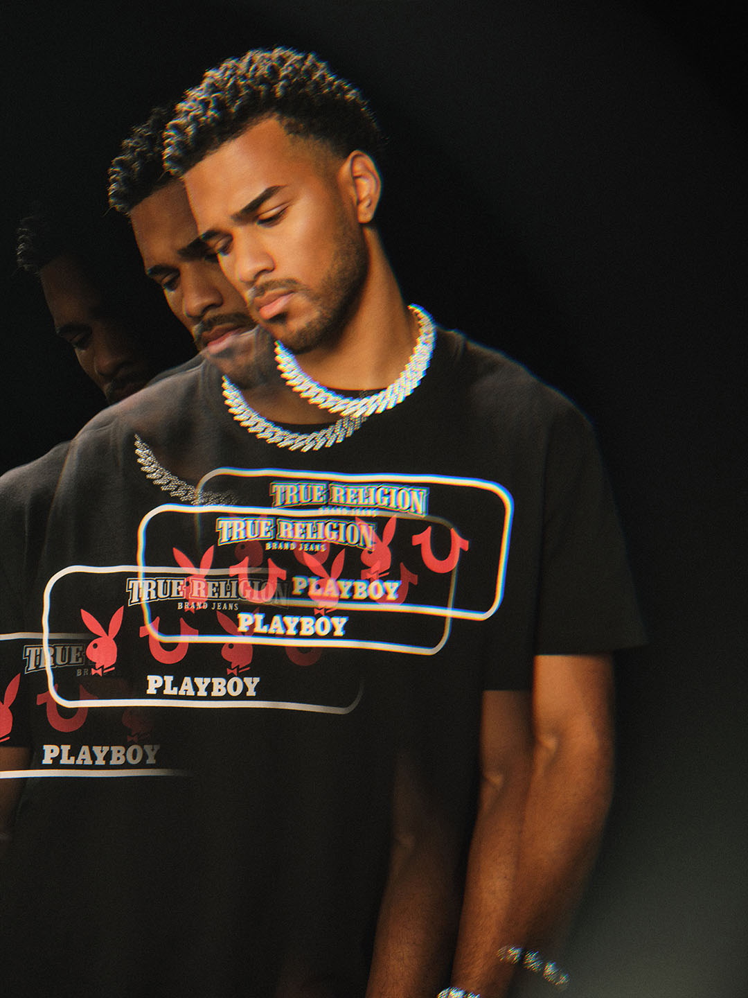 Model wearing a black T-shirt featuring True Religion and Playboy collaboration branding with layered graphic text and red Playboy bunny logos, styled with a silver chain necklace, and presented with a reflective visual effect against a dark background.