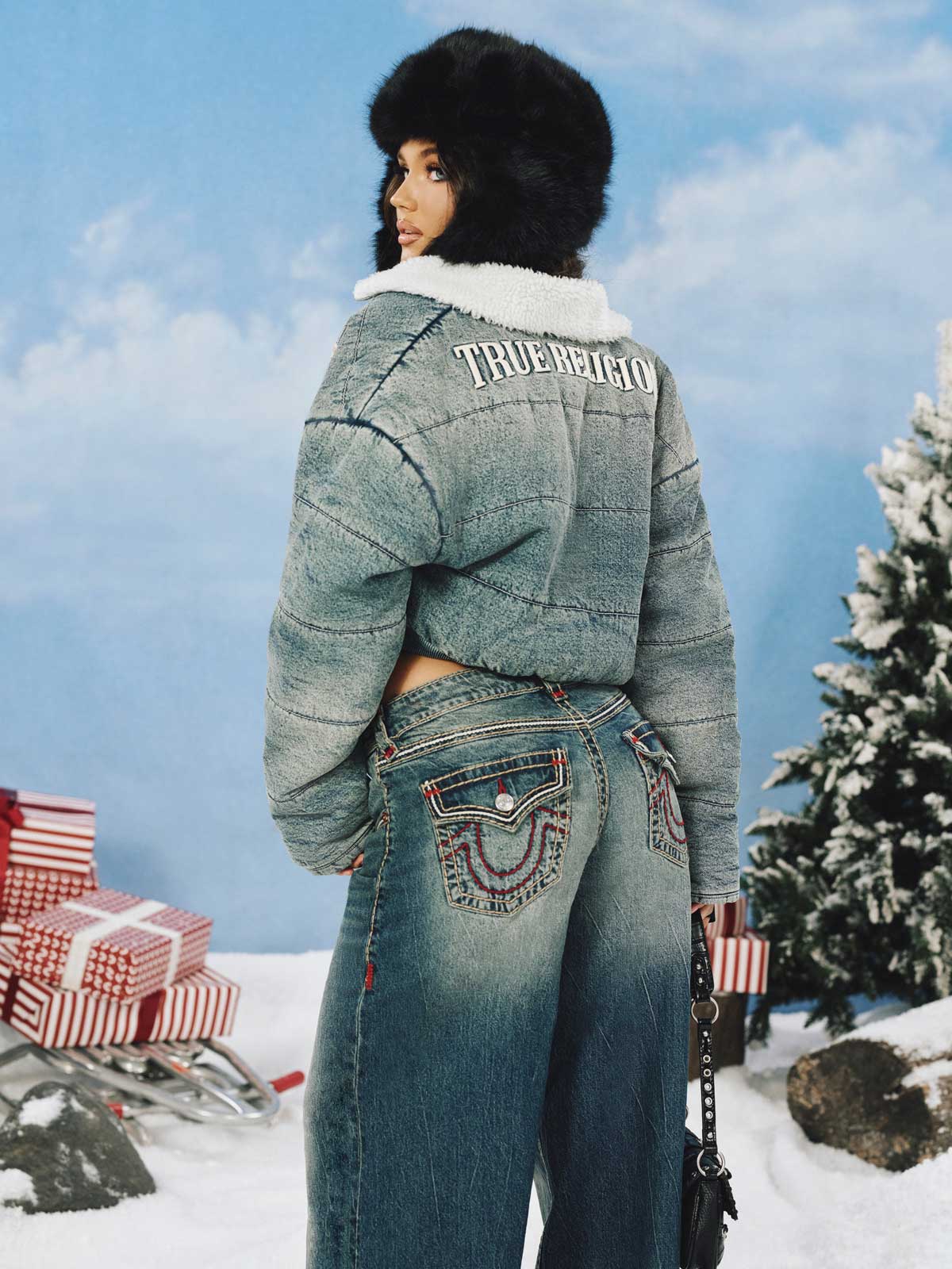 Woman standing in front of an outdoor wintry background wearing a fur hat, a cropped jacket, and barrel jeans 