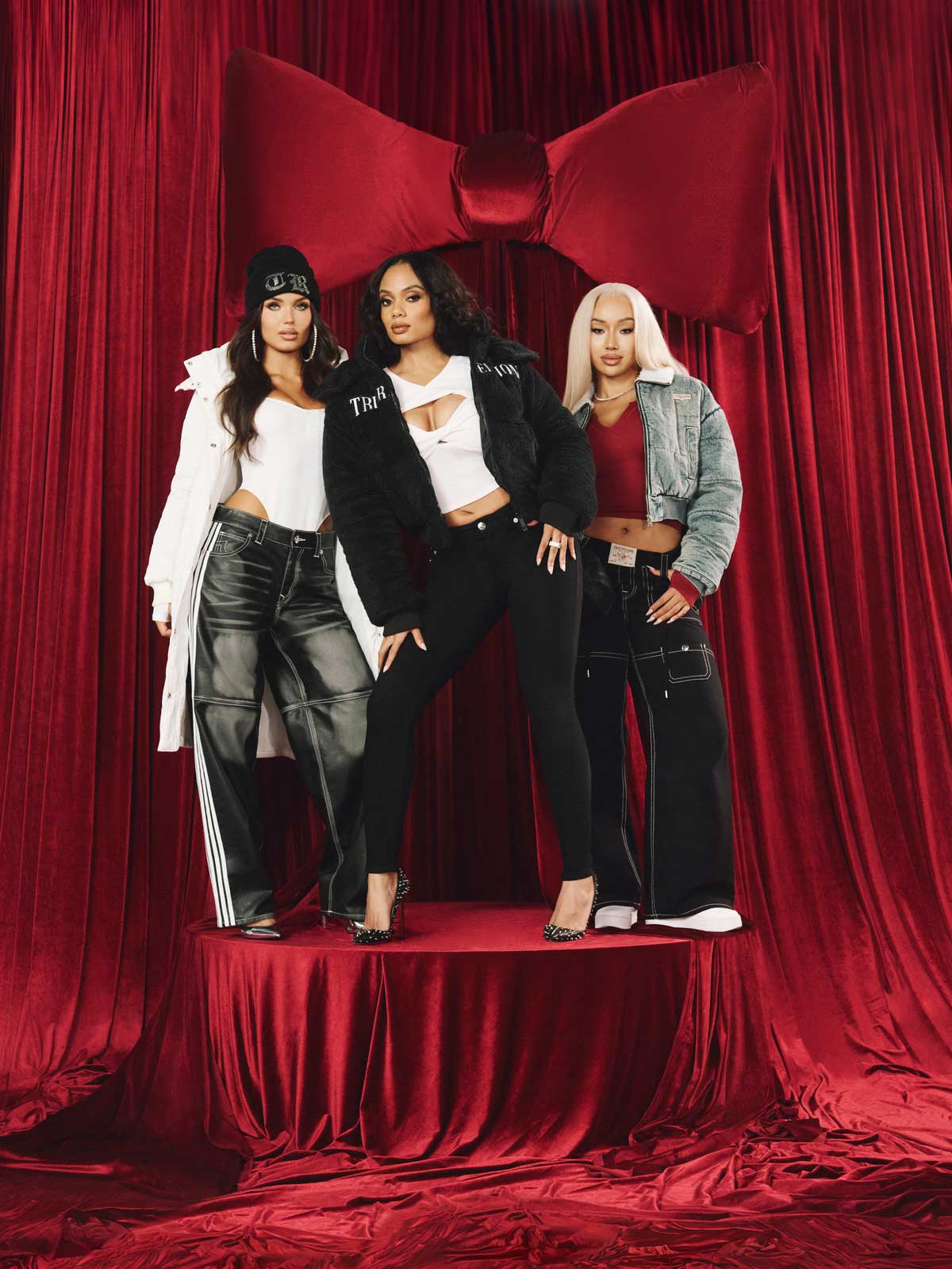 three women stand on a red velvet table. One wears a true religion white puffer jacket and acid wash jeans, another wears a true religion black puffer jacket and black skinny jeans, another wears a cropped gray jacket and wide leg jeans