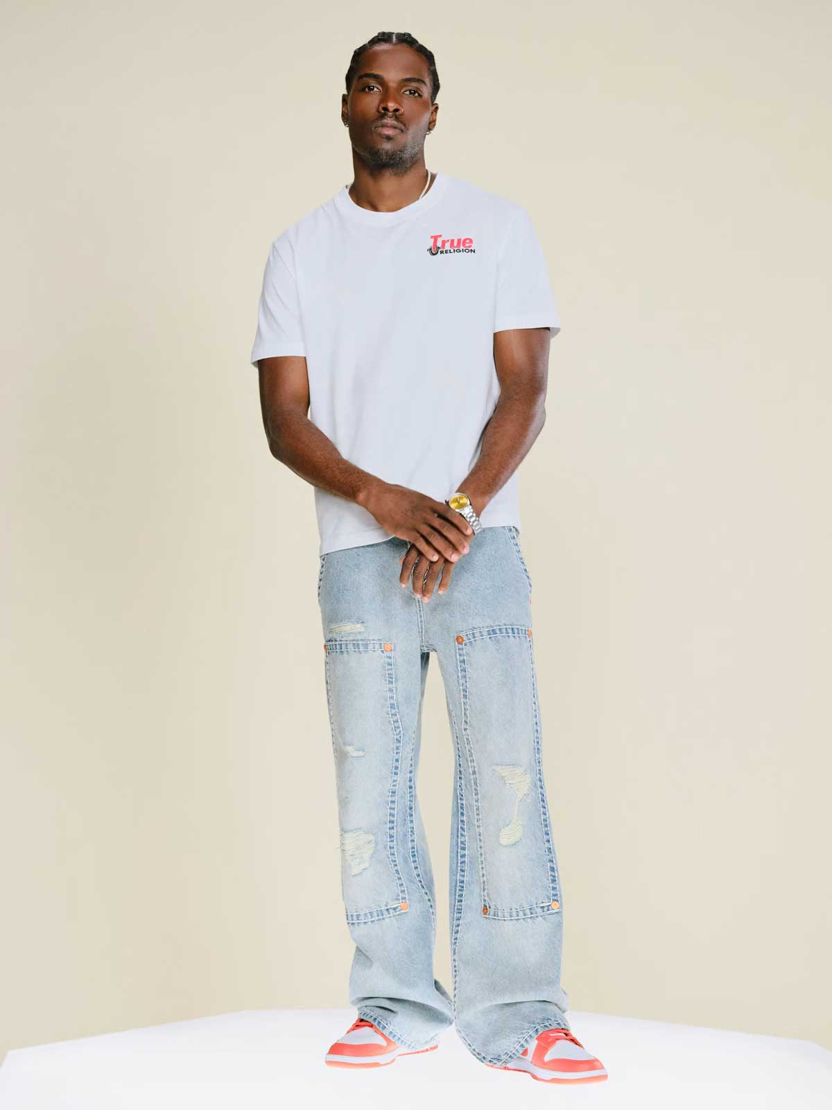 Man wears true religion white tee shirt and true religion light wash baggy jeans with patch details.
