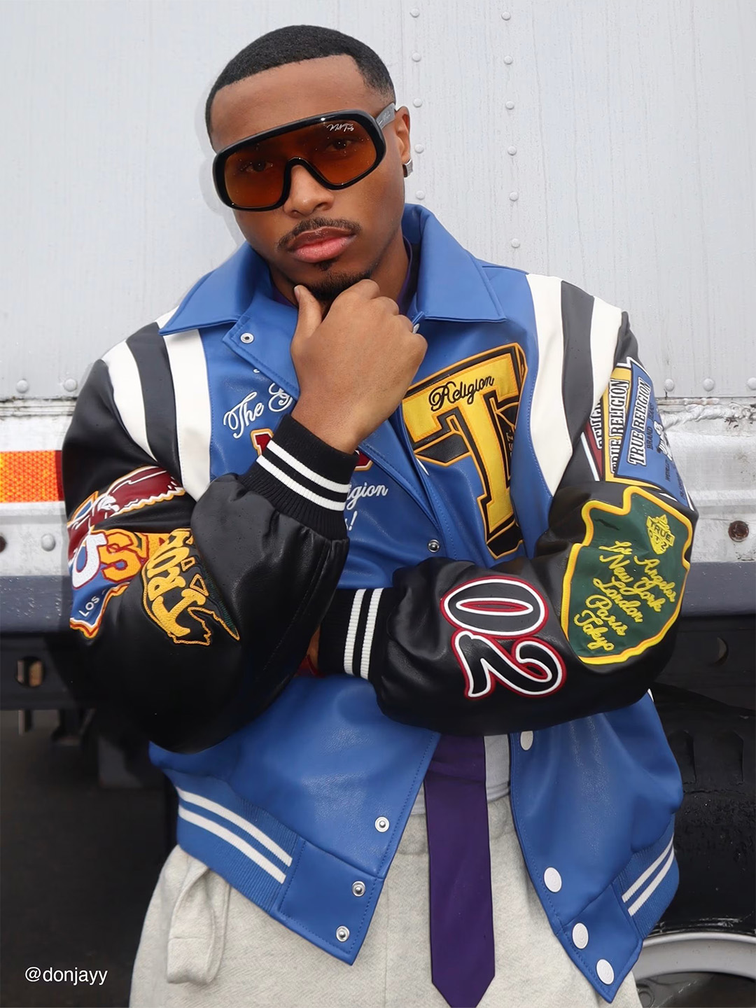 Man wears black sunglasses and a blue, black, white and yellow true religion varsity jacket.