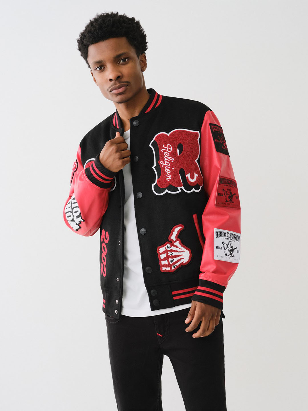 Man posing wearing a black true religion varsity jacket with red and white sleeves and patches, and dark wash true religion jeans.